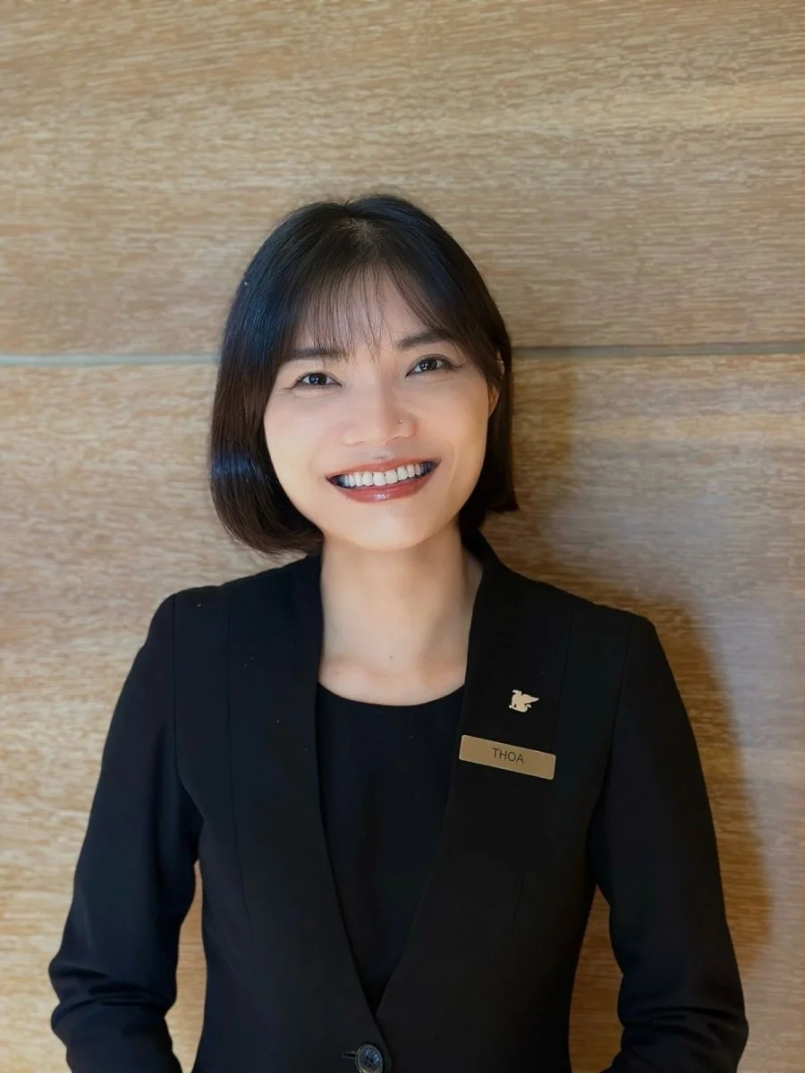 A woman with short black hair and a black blazer smiling, standing against a wooden wall.