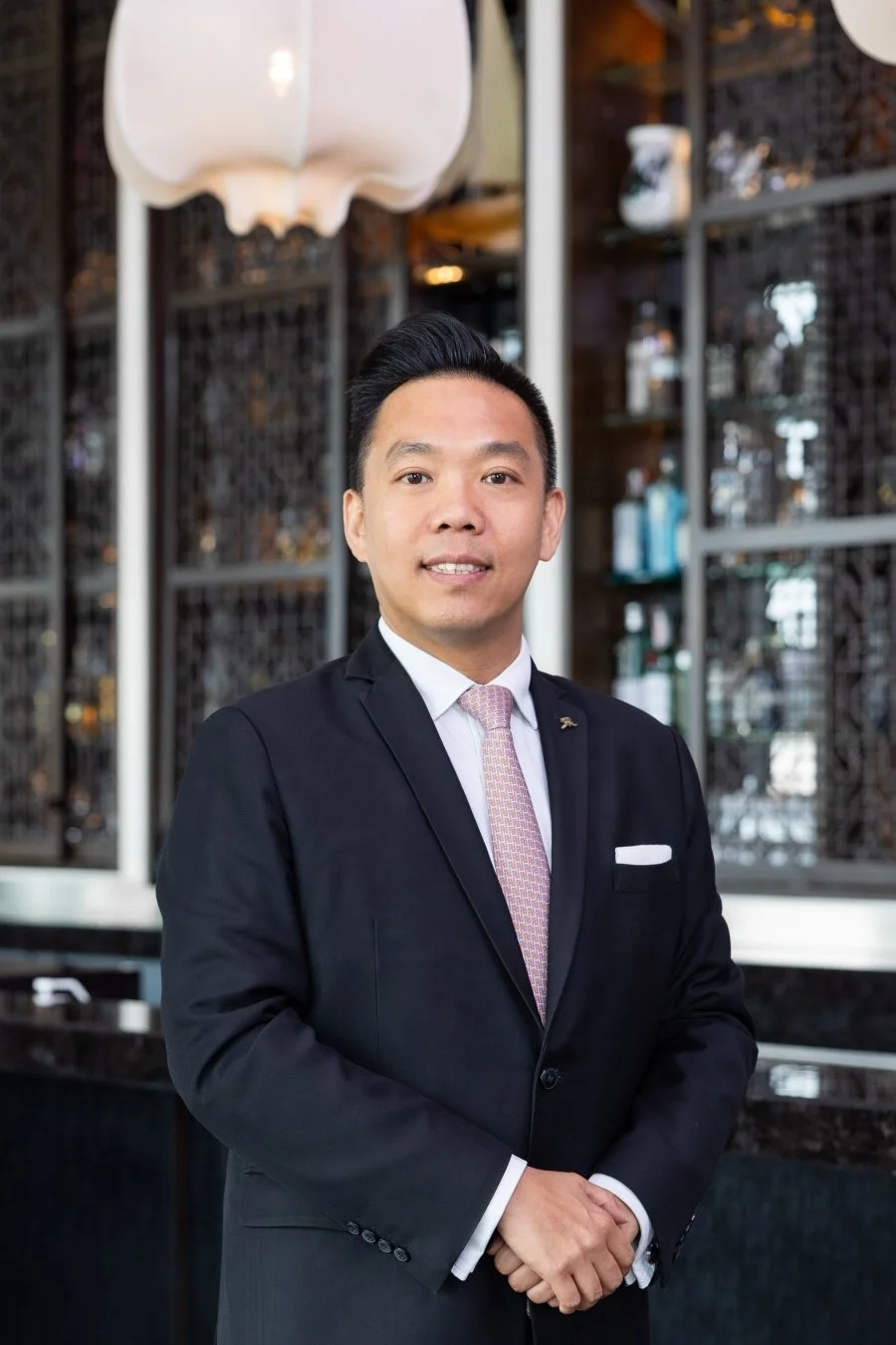 A man in a black suit and pink tie standing indoors in front of a dark decorative wall with shelves and bottles.