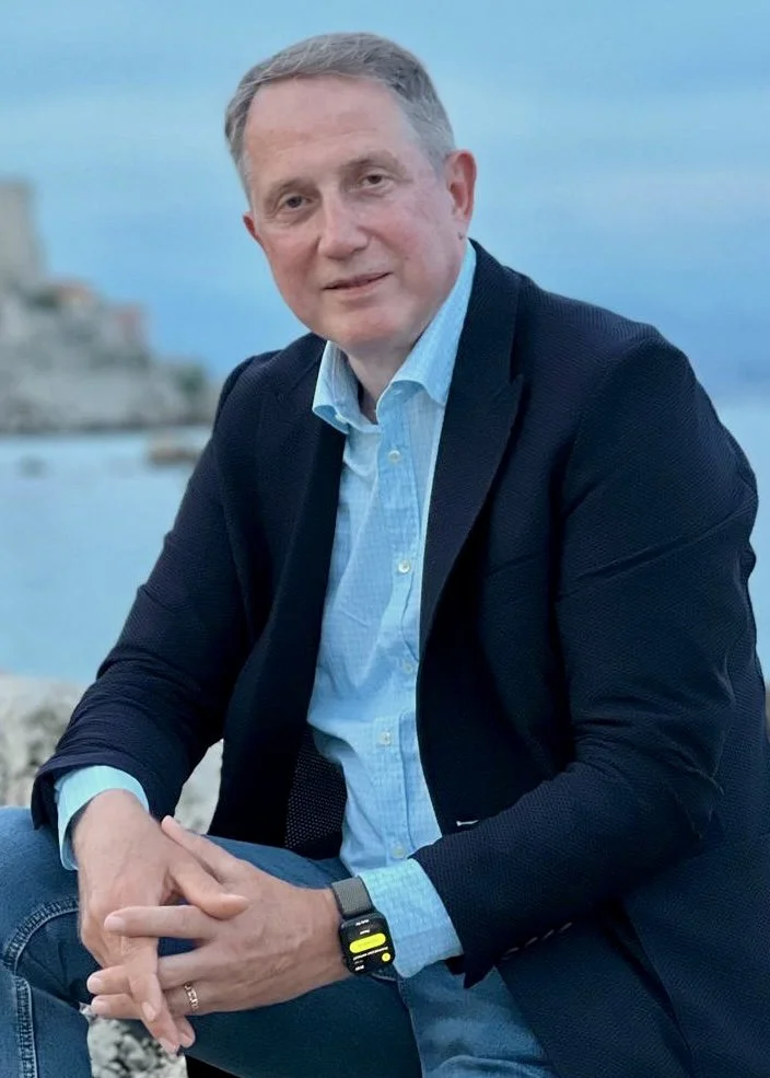 A man in a checked brown blazer and blue shirt with a blurred green background.