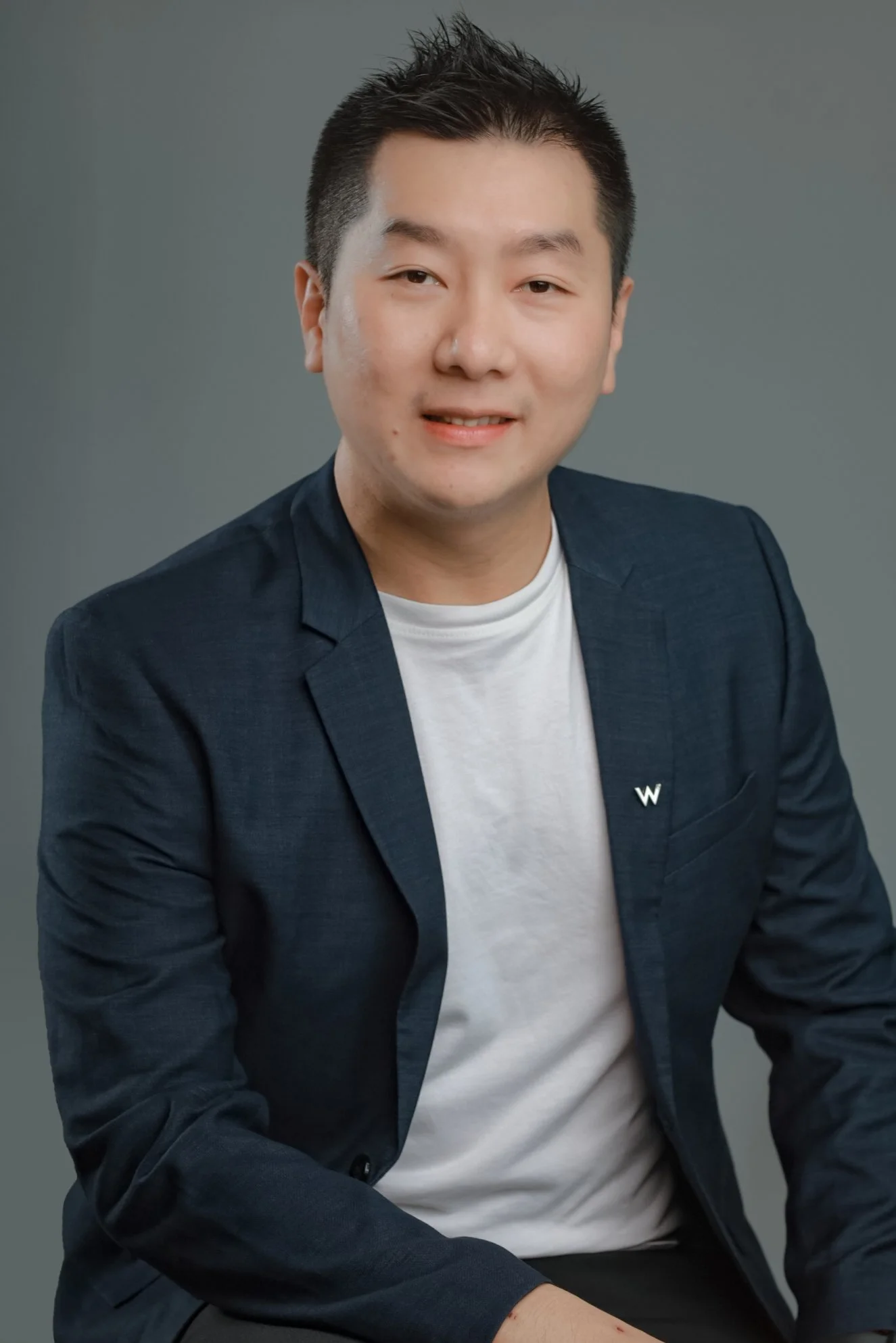 Portrait of a young Asian man with black hair, smiling, dressed in a navy blazer and white t-shirt, sitting with arms crossed on a table, wearing a smartwatch, against a gray background.