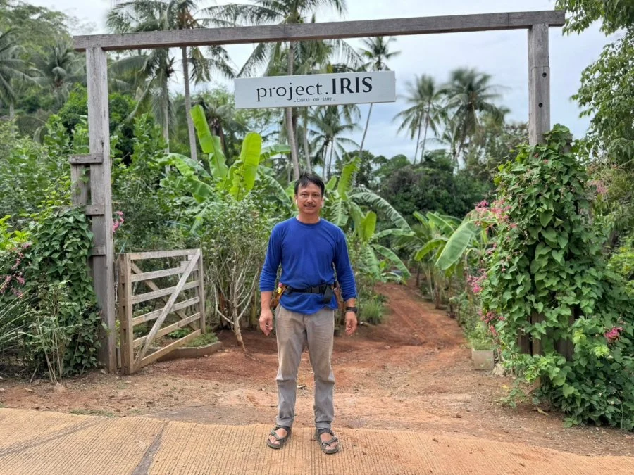A man standing on a dirt path beneath a sign that reads 'project.IRIS' surrounded by lush green tropical plants and banana trees.