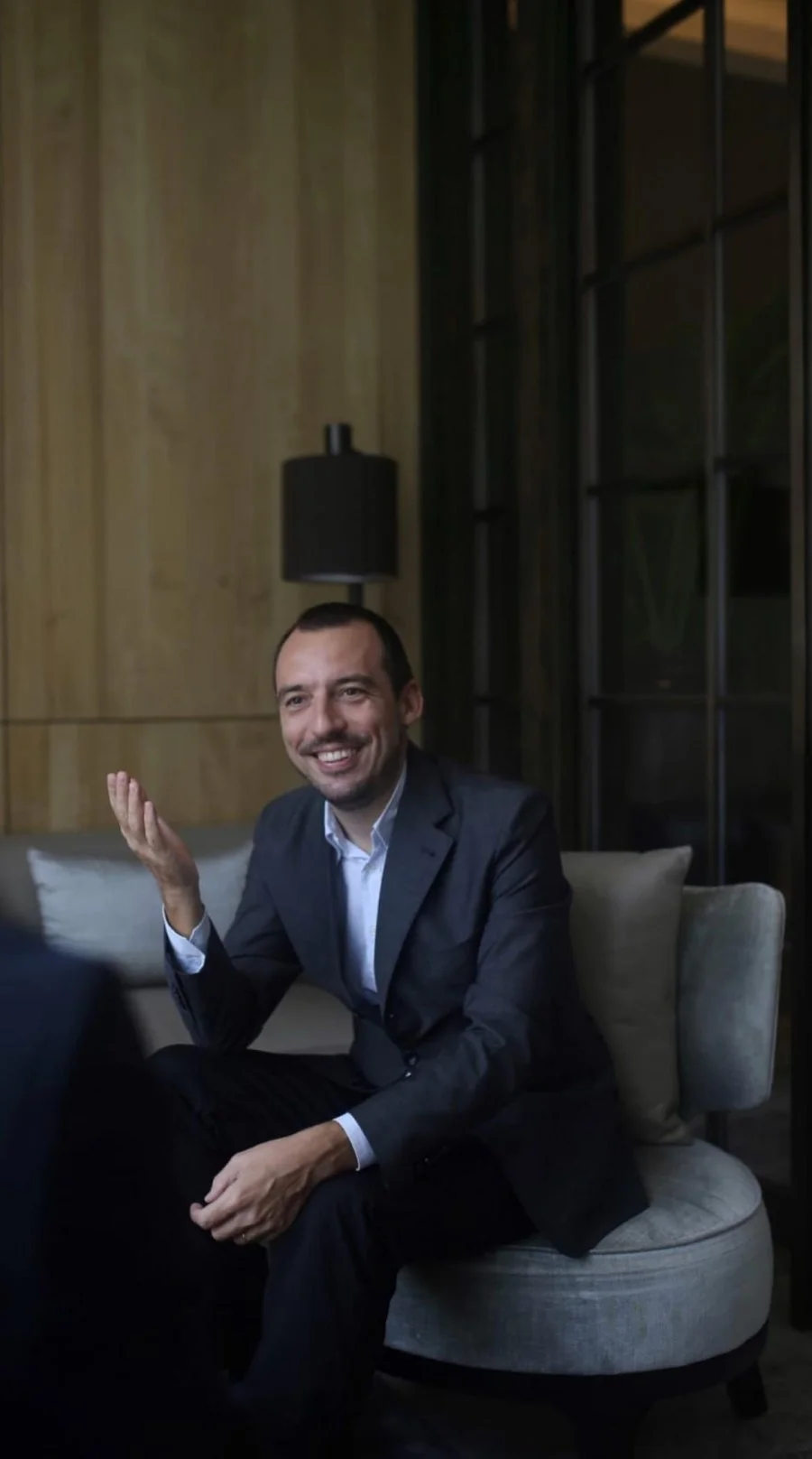A man in a gray suit and white shirt sitting on a cream-colored sofa in a modern, dimly lit interior, smiling and gesturing as if engaged in conversation.