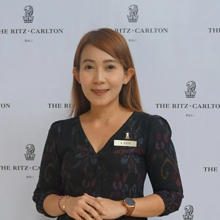 Woman with shoulder-length brown hair, wearing a black floral dress, a name tag labeled 'Lady,' a bracelet, a watch, and a ring, standing in front of a backdrop with 'The Ritz-Carlton Bali' logos.