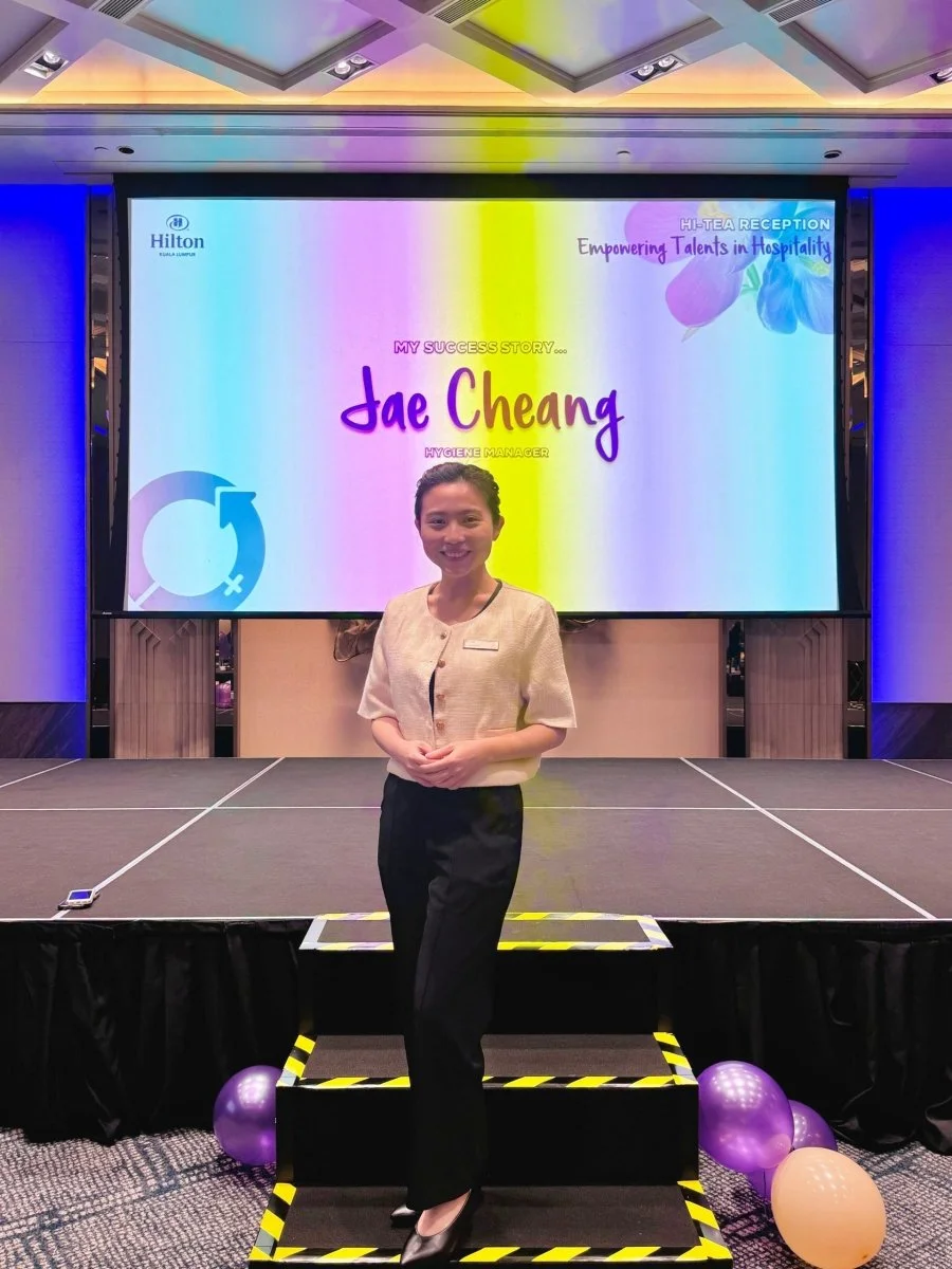 A woman standing on a small stage with yellow and black caution tape borders, in front of a large screen displaying a presentation slide. The slide shows the Hilton logo, text that says "My Success Story... Jae Cheang, Hygiene Manager," and a colorful background with flowers and a gender symbol. There are purple and orange balloons on the floor near the stage.