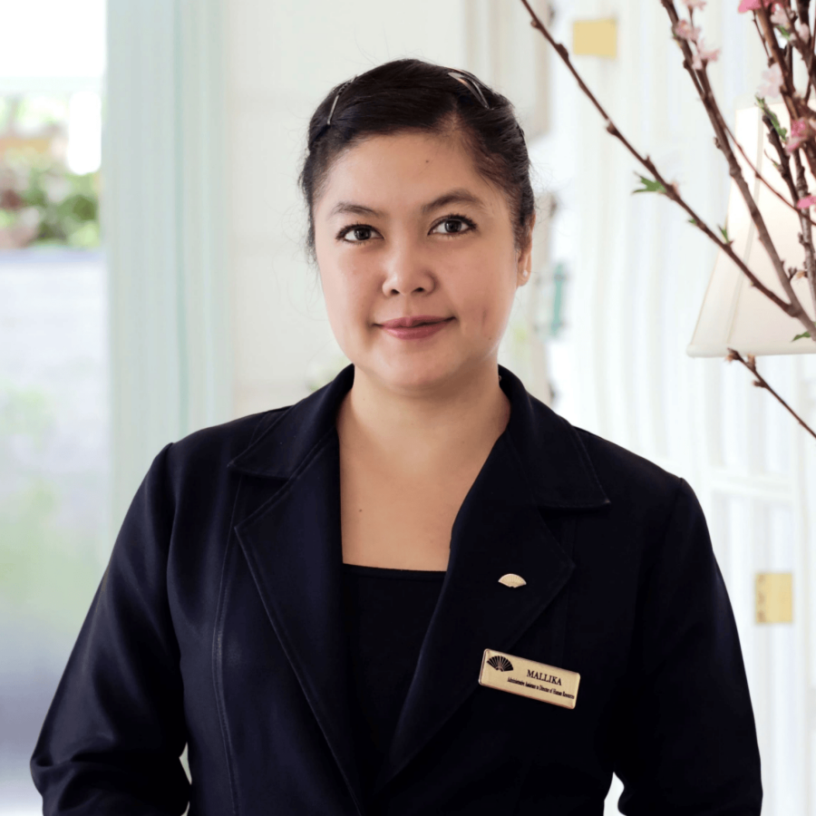 A woman with dark hair styled back, wearing a black uniform with a name tag that says 'Mallika,' stands indoors next to a branch with pink blossoms, against a light-colored background.