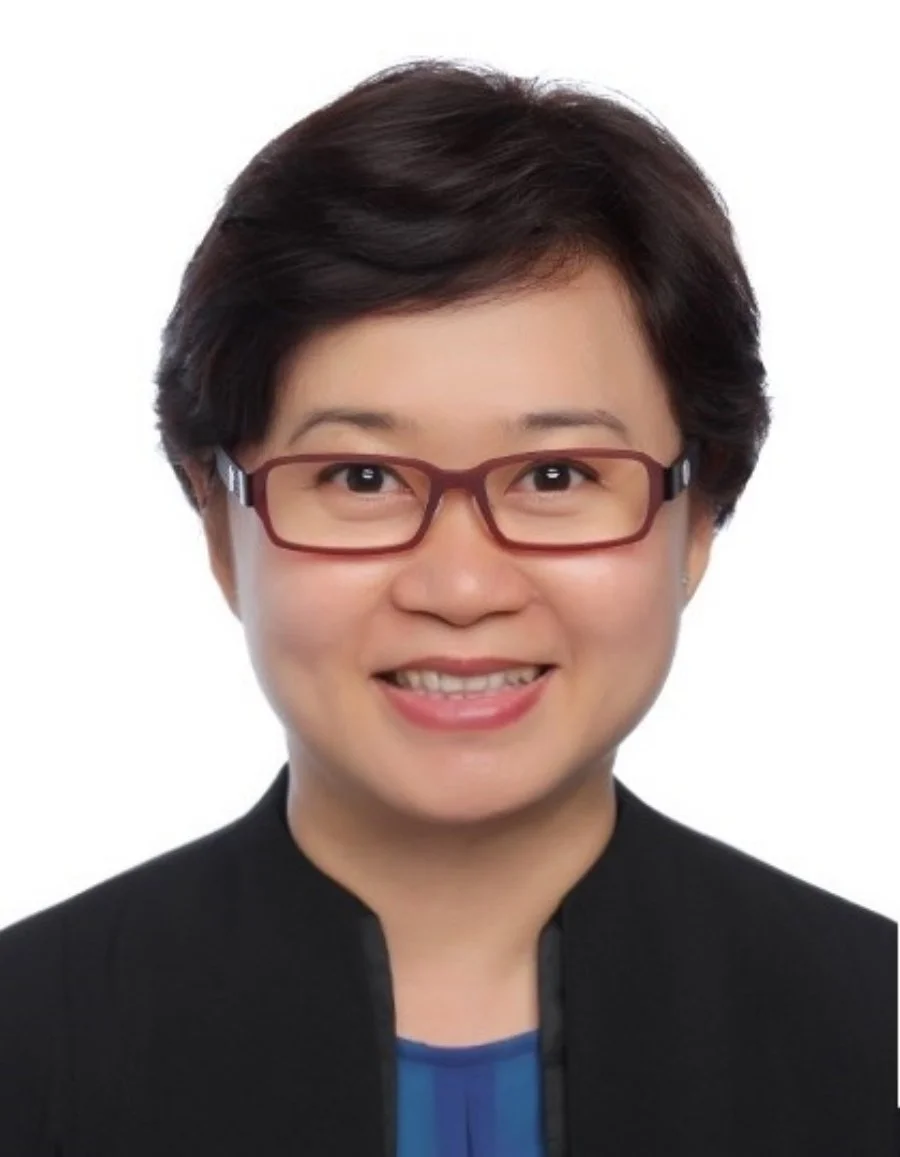 Headshot of an Asian woman with short dark hair, wearing glasses, a black jacket, and a blue top, smiling against a white background.