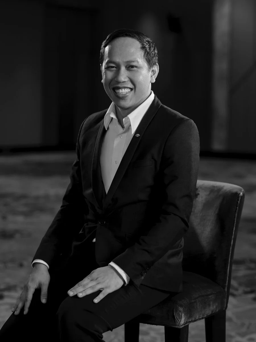 A black and white portrait of a smiling man in a suit sitting on a chair in an indoor setting.