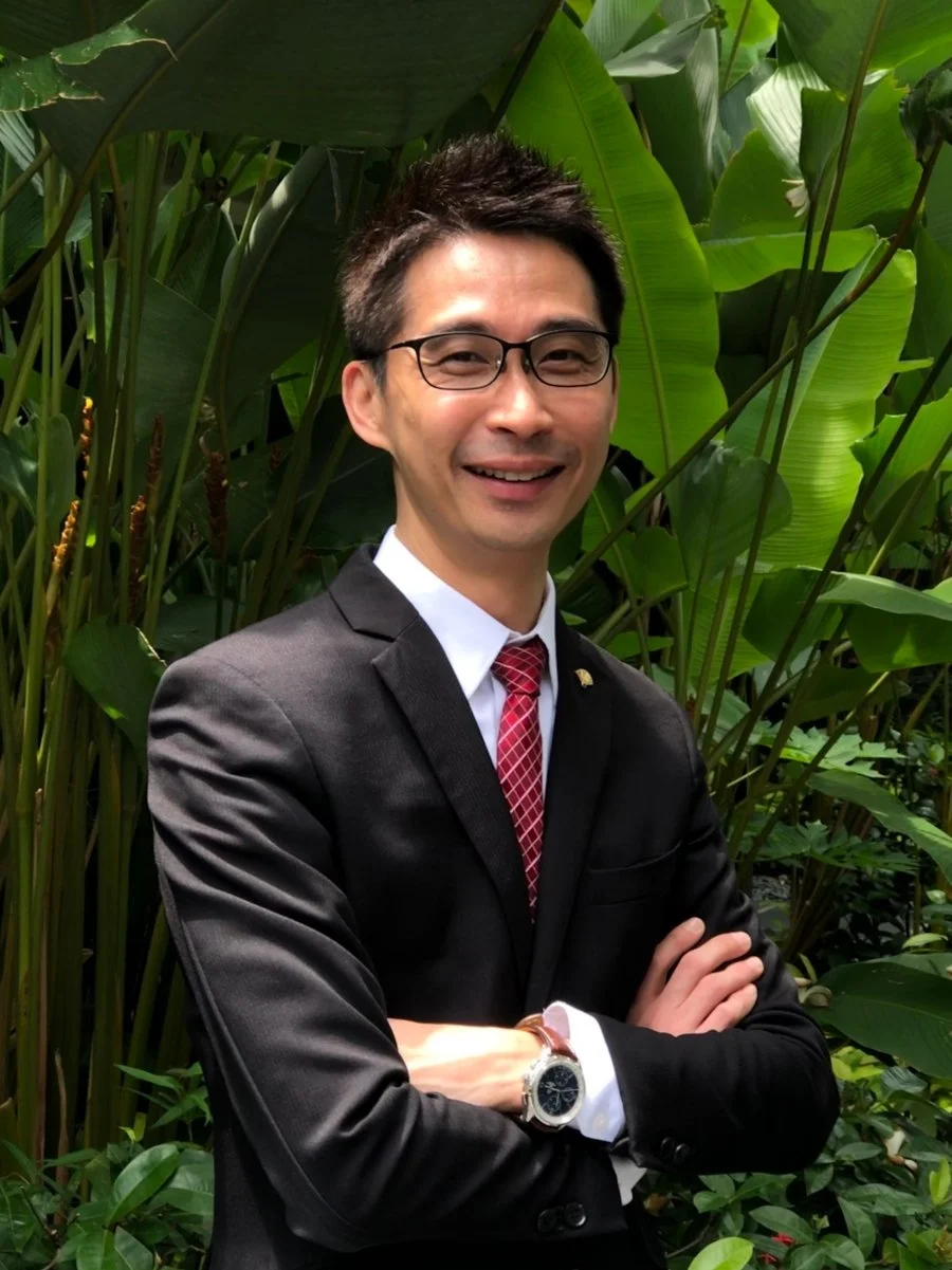 A man in a black suit, white shirt, and red tie stands outdoors in front of lush green tropical plants, smiling with arms crossed, wearing glasses and a wristwatch.