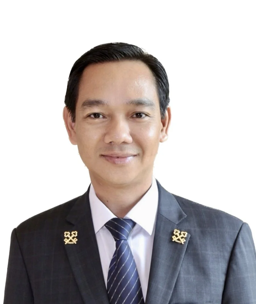 A man in a dark suit, white shirt, and striped tie, smiling, with a plain white background.