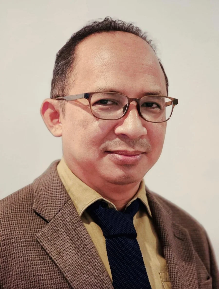 A middle-aged man with glasses, short dark hair, wearing a brown blazer, tan shirt, and dark tie, standing against a plain light background.