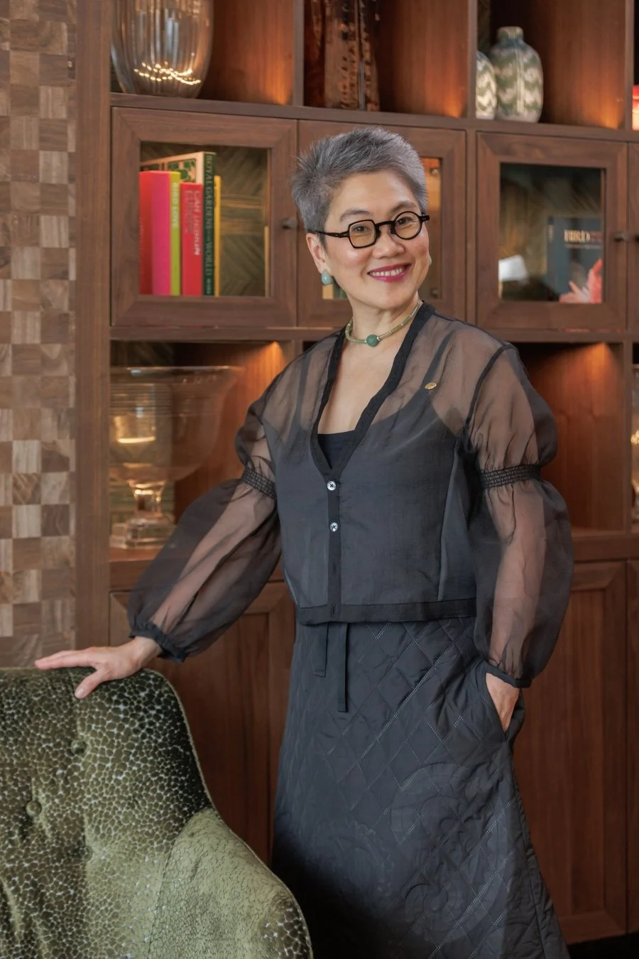 A smiling woman with short gray hair, glasses, and turquoise jewelry standing in front of a wooden bookshelf filled with books and decorative objects, wearing a black sheer top with puffed sleeves and a quilted skirt.