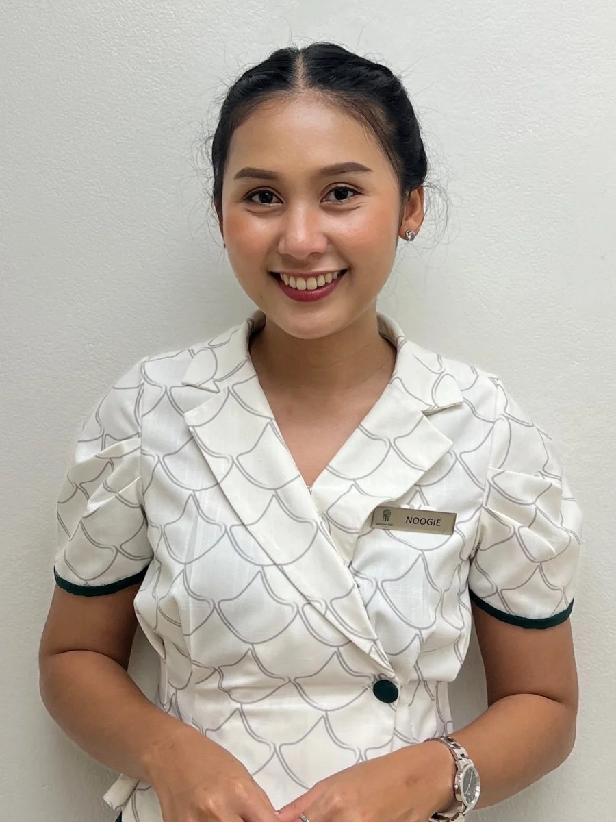 A young woman with dark hair in a braid, wearing earrings, smiling, in a white patterned uniform with a name tag that reads 'Noogie,' standing against a plain white wall.