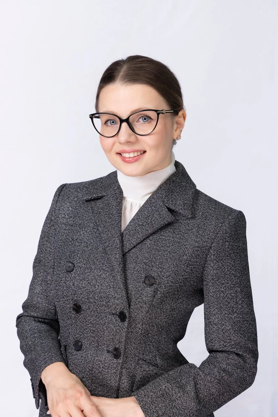 Professional woman with glasses, dark hair in a bun, wearing a gray double-breasted blazer and a white turtleneck, smiling against a plain white background.