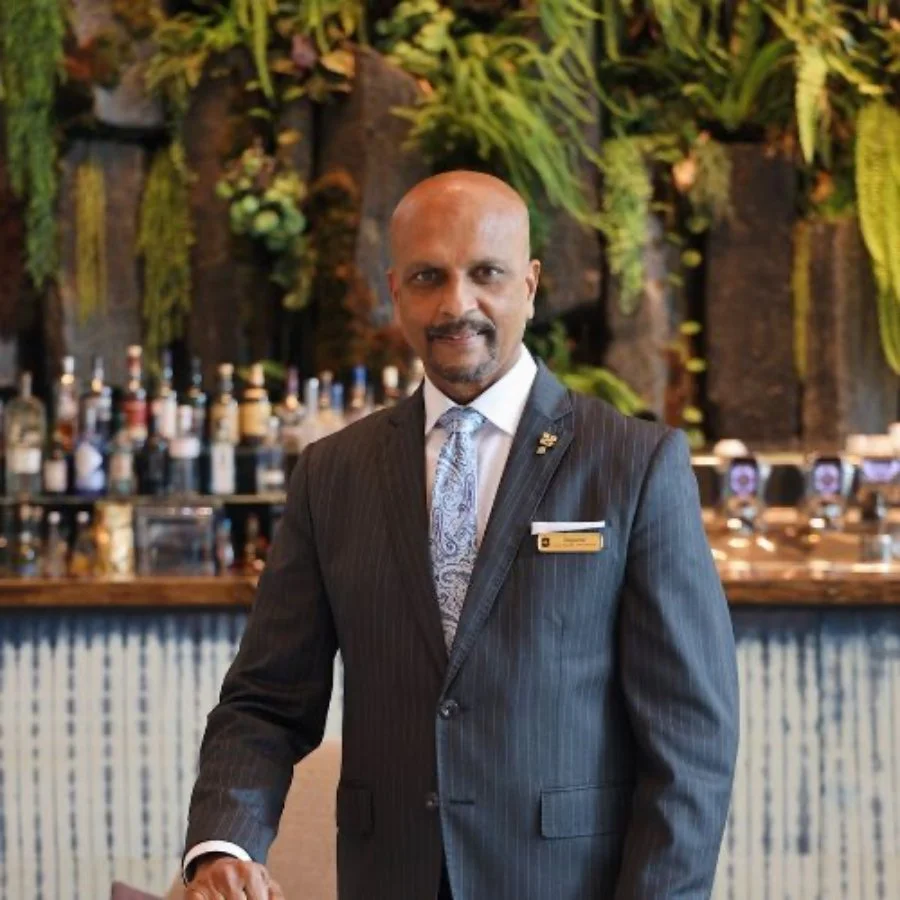 A man in a dark pinstripe suit and patterned tie standing in front of a bar with bottles and greenery in the background.