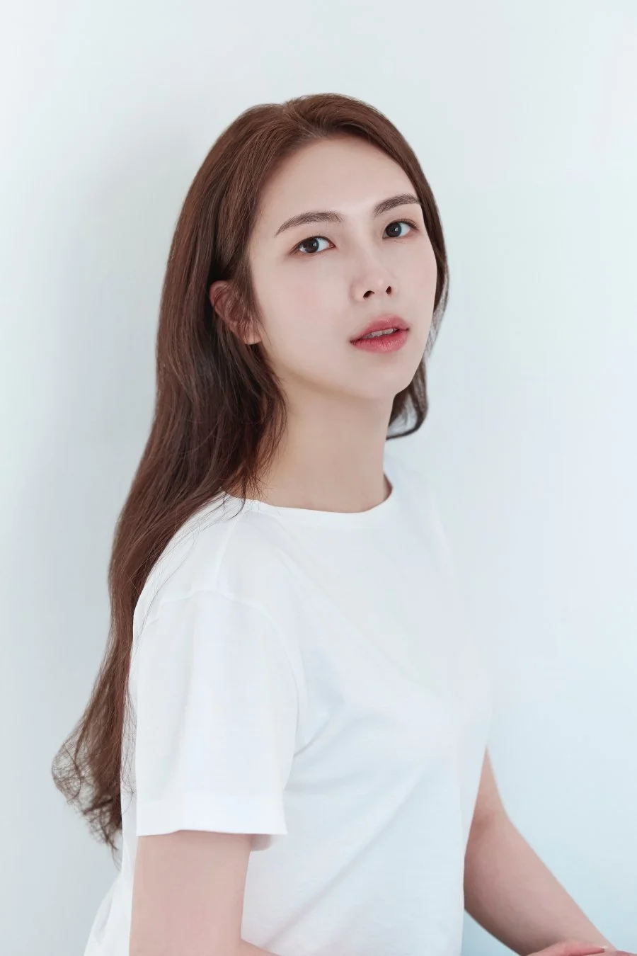 A young woman with long brown hair, light skin, and wearing a white t-shirt, looks at the camera against a plain light background.