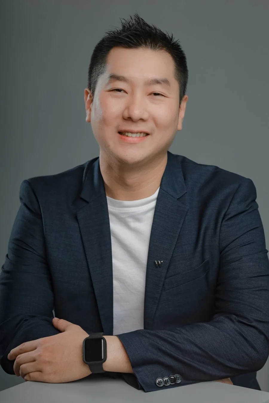 Portrait of a young Asian man with black hair, smiling, dressed in a navy blazer and white t-shirt, sitting with arms crossed on a table, wearing a smartwatch, against a gray background.