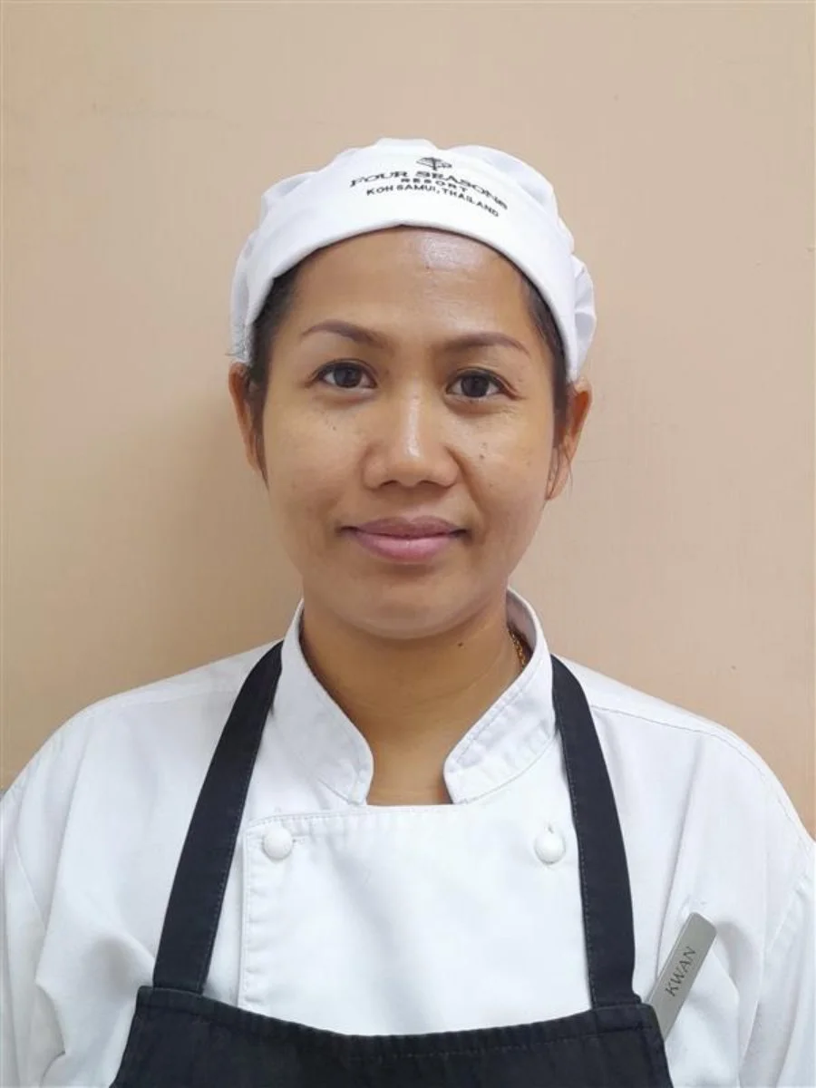 Female chef in white uniform and apron, wearing a white headscarf with writing, standing against a neutral background.