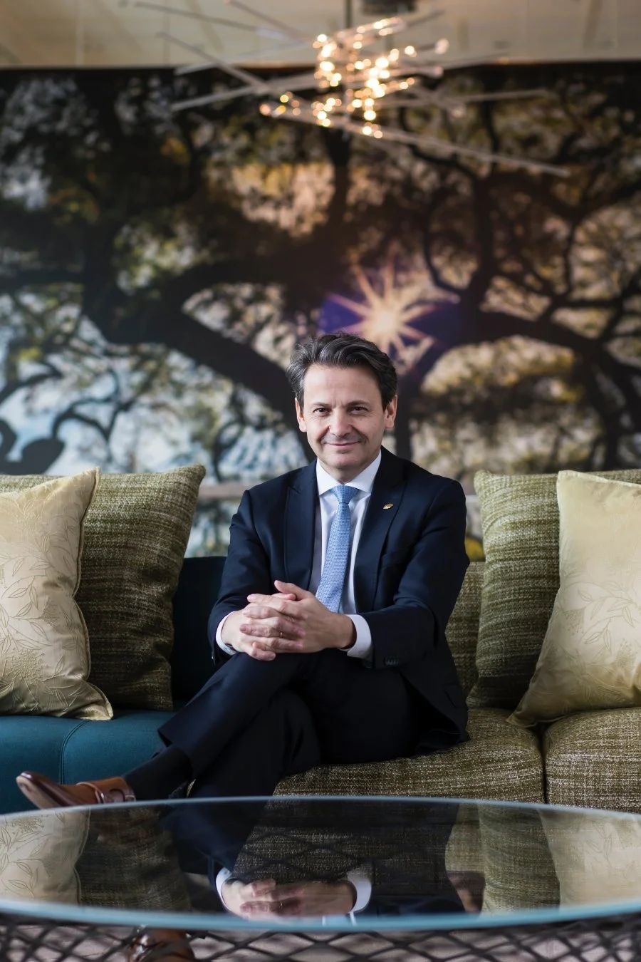 A man in a dark suit with a light blue tie sitting on a sofa in a luxurious modern interior with a large mural of trees and a sunset in the background, and a glass coffee table in the foreground.