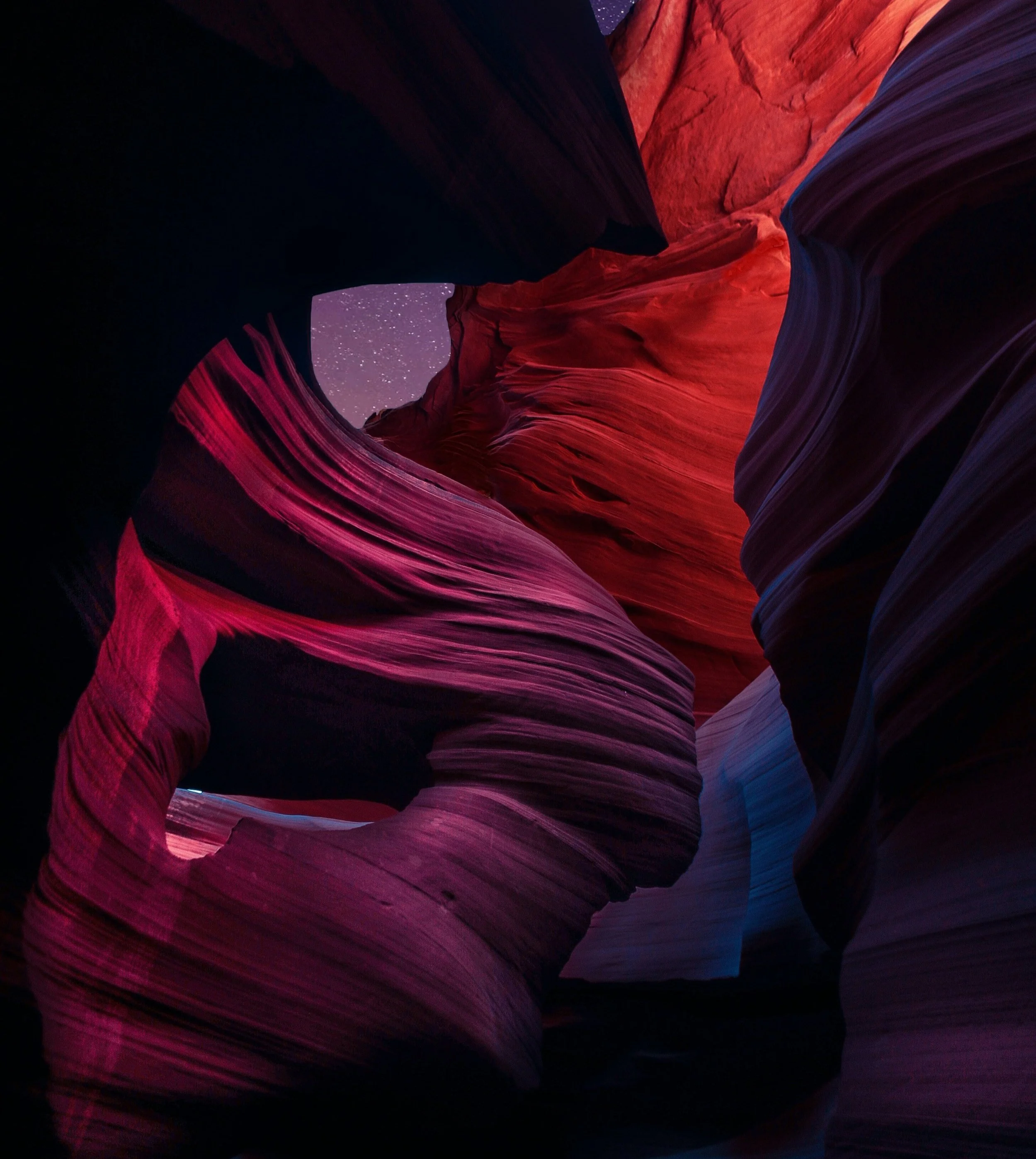 A colorful slot canyon with smooth, flowing rock formations in red, purple, and orange hues, sky with stars visible through opening
