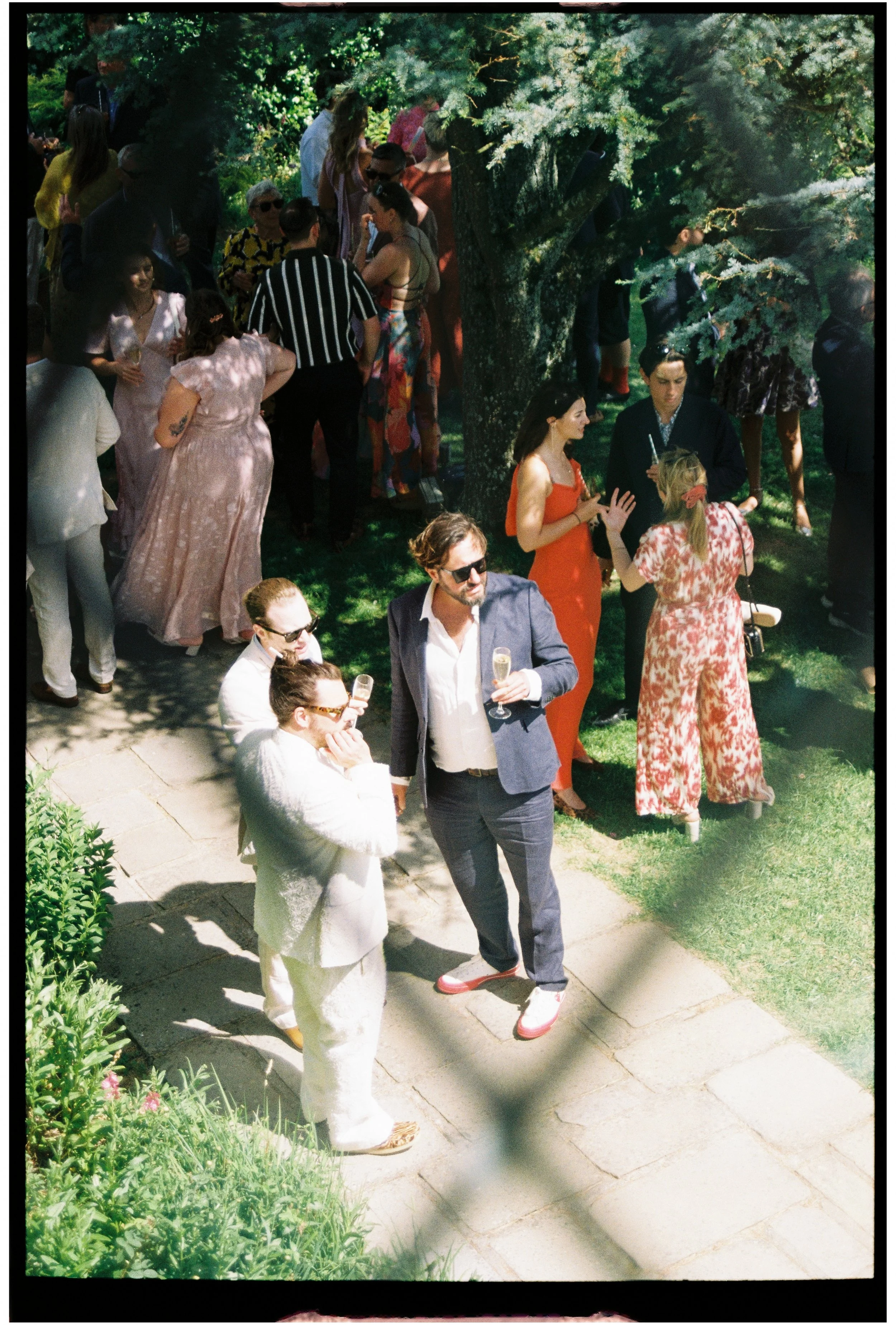 A group of people gathered outdoors on a sunny day, socializing among trees and greenery, some holding drinks, dressed in semi-formal attire, with a mix of men and women engaged in conversations.