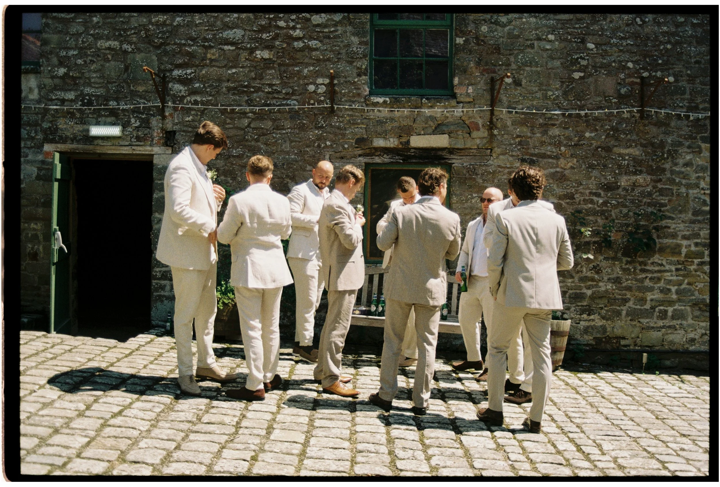 A group of men dressed in beige suits gathers outdoors in front of a stone building, some holding drinks, during daytime.