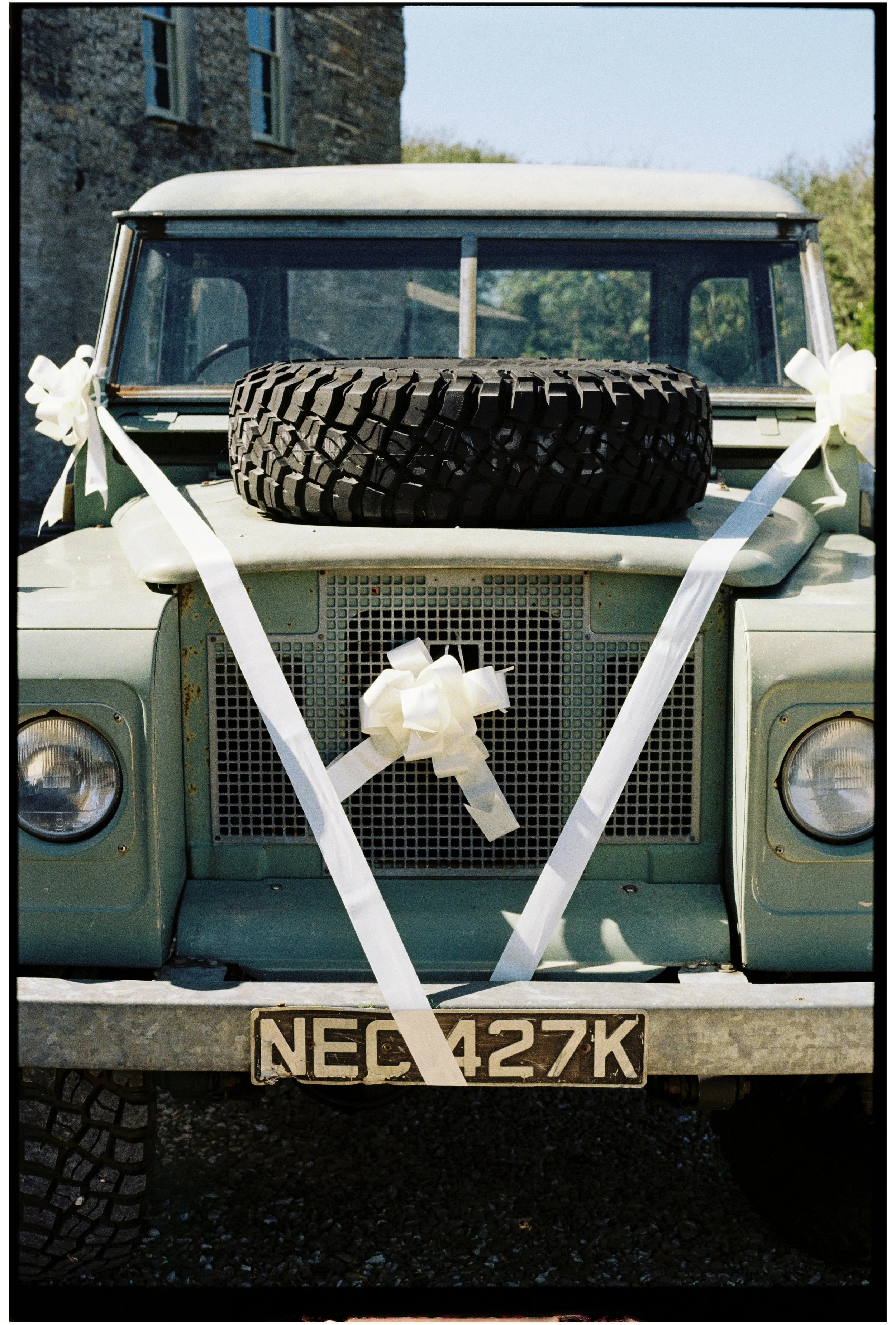 Front of an old beige Land Rover with decorative white ribbons and bows attached, a spare tire on the hood, a visible license plate reading 'NEC 427K', and an old stone building and trees in the background.