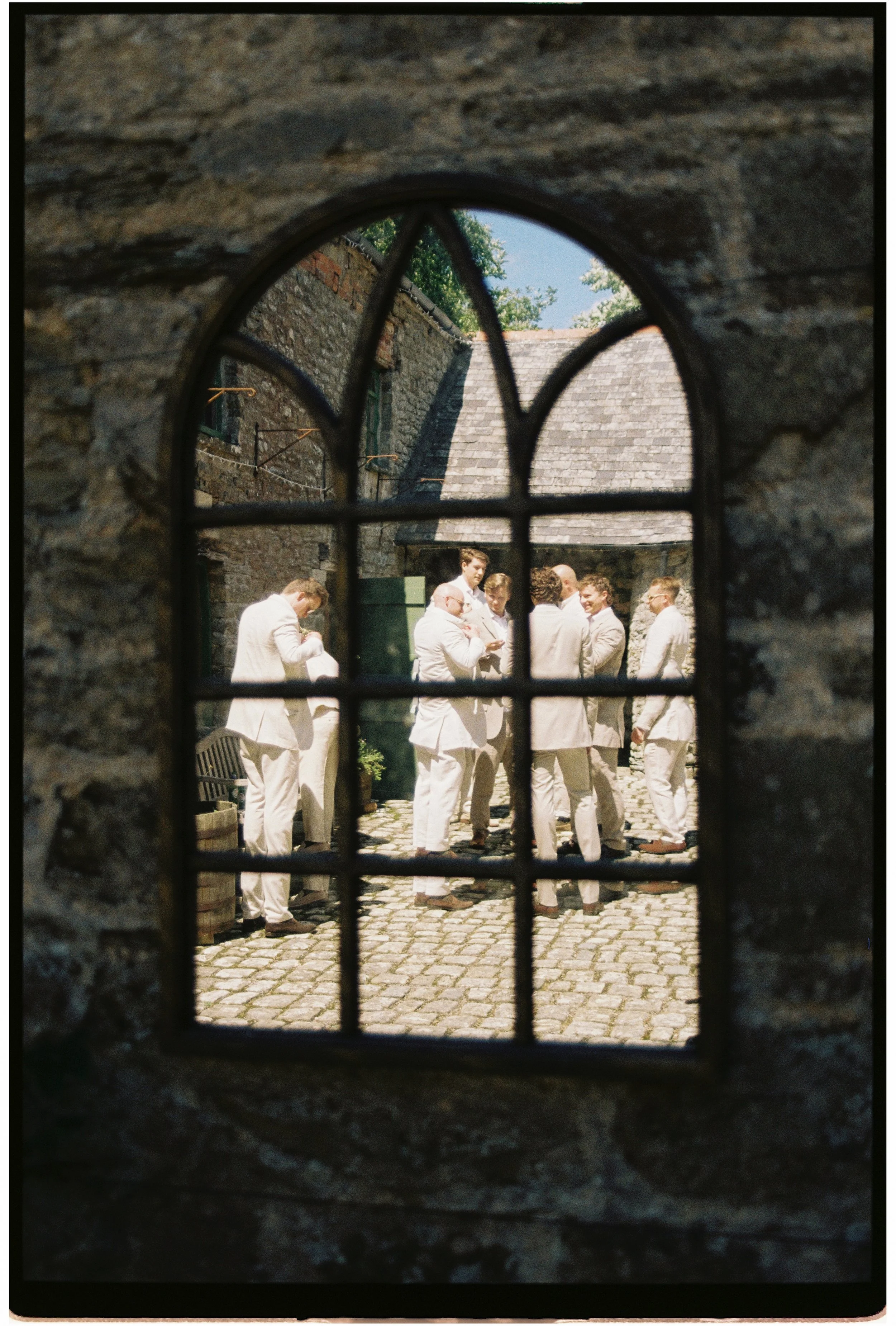 View through a small, iron-barred mirror of a group of ushers in white suits gathered for the wedding day in a cobblestone courtyard outside of the old stone building at Ash Barton Estate, Devon.