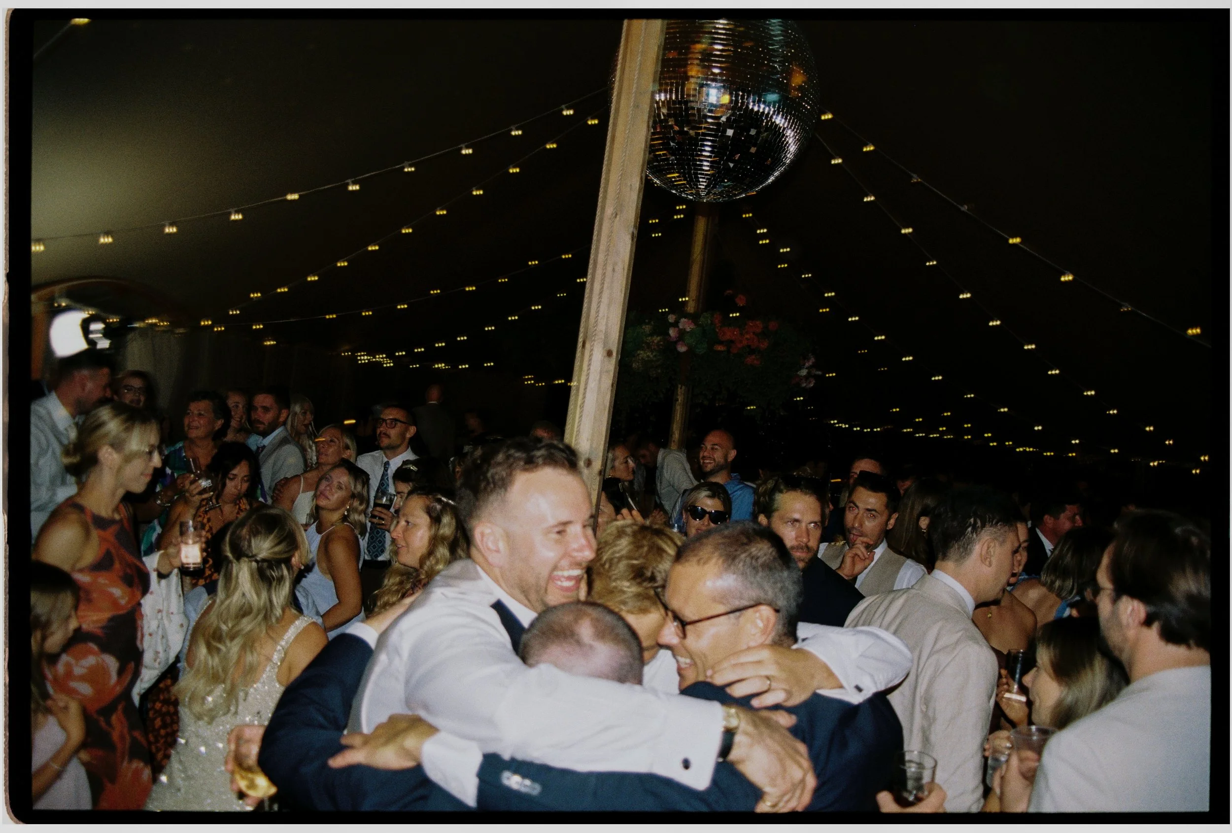 Crowd of people at a lively party or celebration with string lights, a disco ball, and floral decorations hanging from the ceiling.