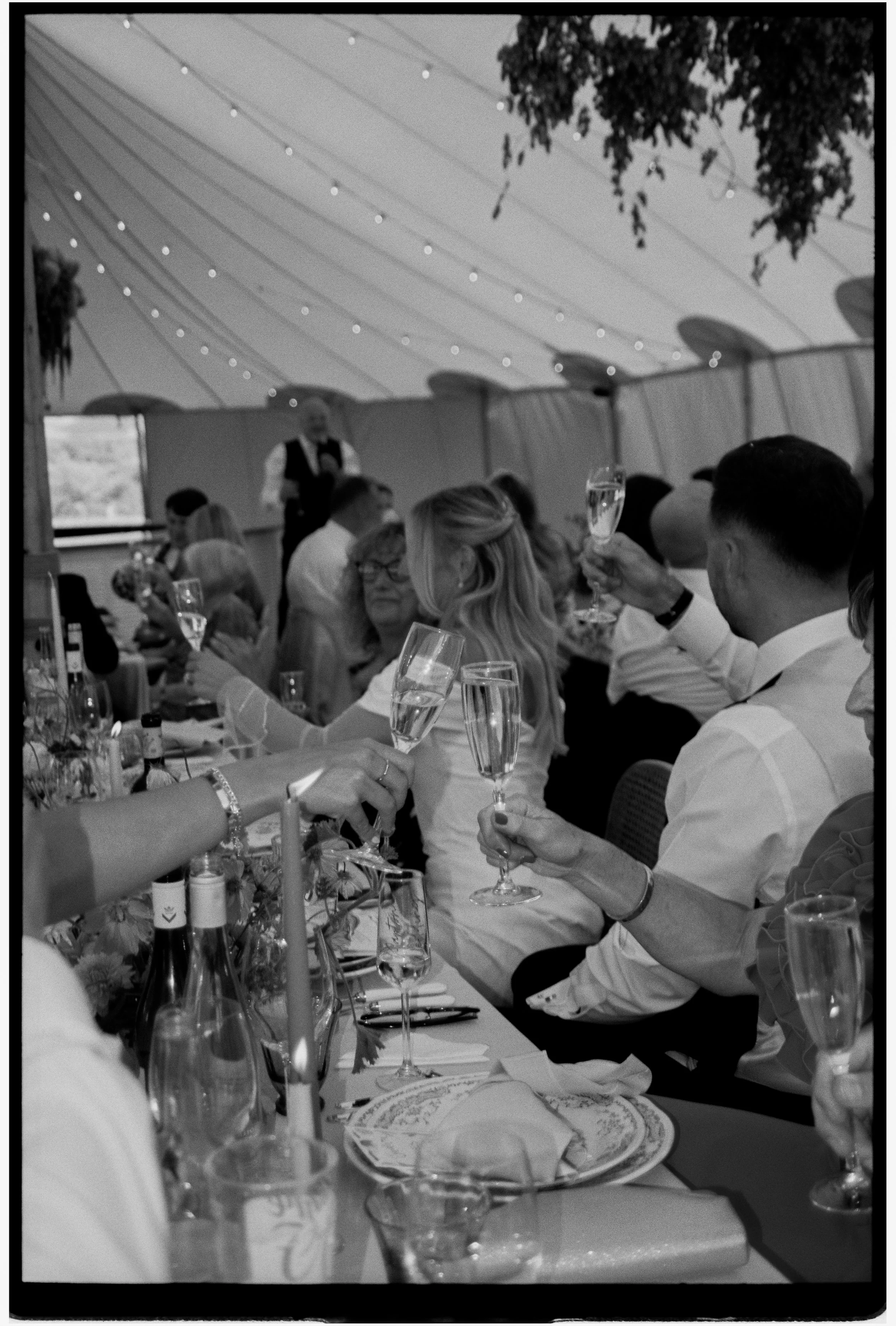 People celebrating at a wedding reception in a decorated tent with string lights, drinking champagne and enjoying a meal.