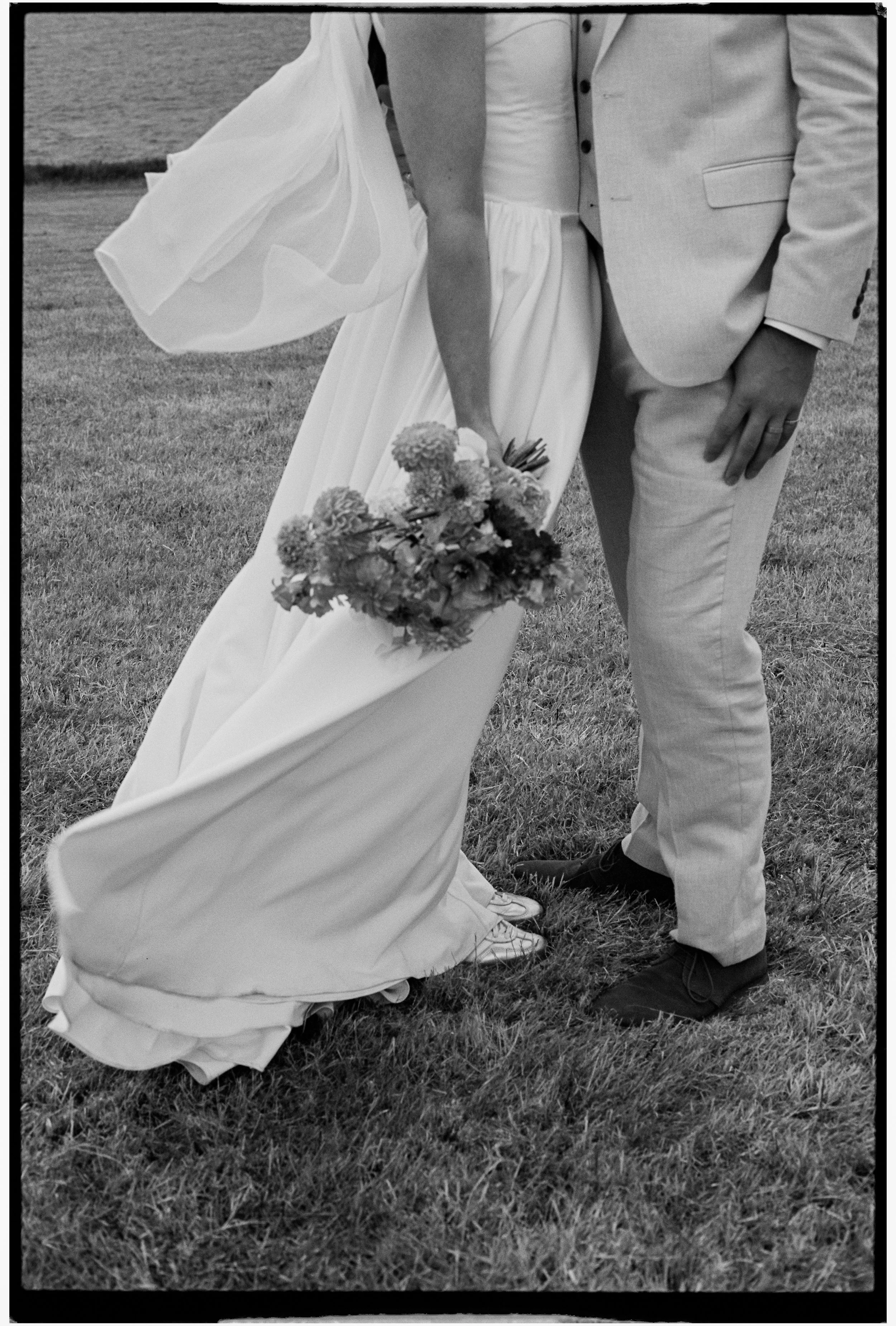 Close-up black and white photo of a bride and groom standing outdoors on grassy land, with the bride holding a bouquet of flowers, both dressed in wedding attire, showing only the lower half of their bodies.