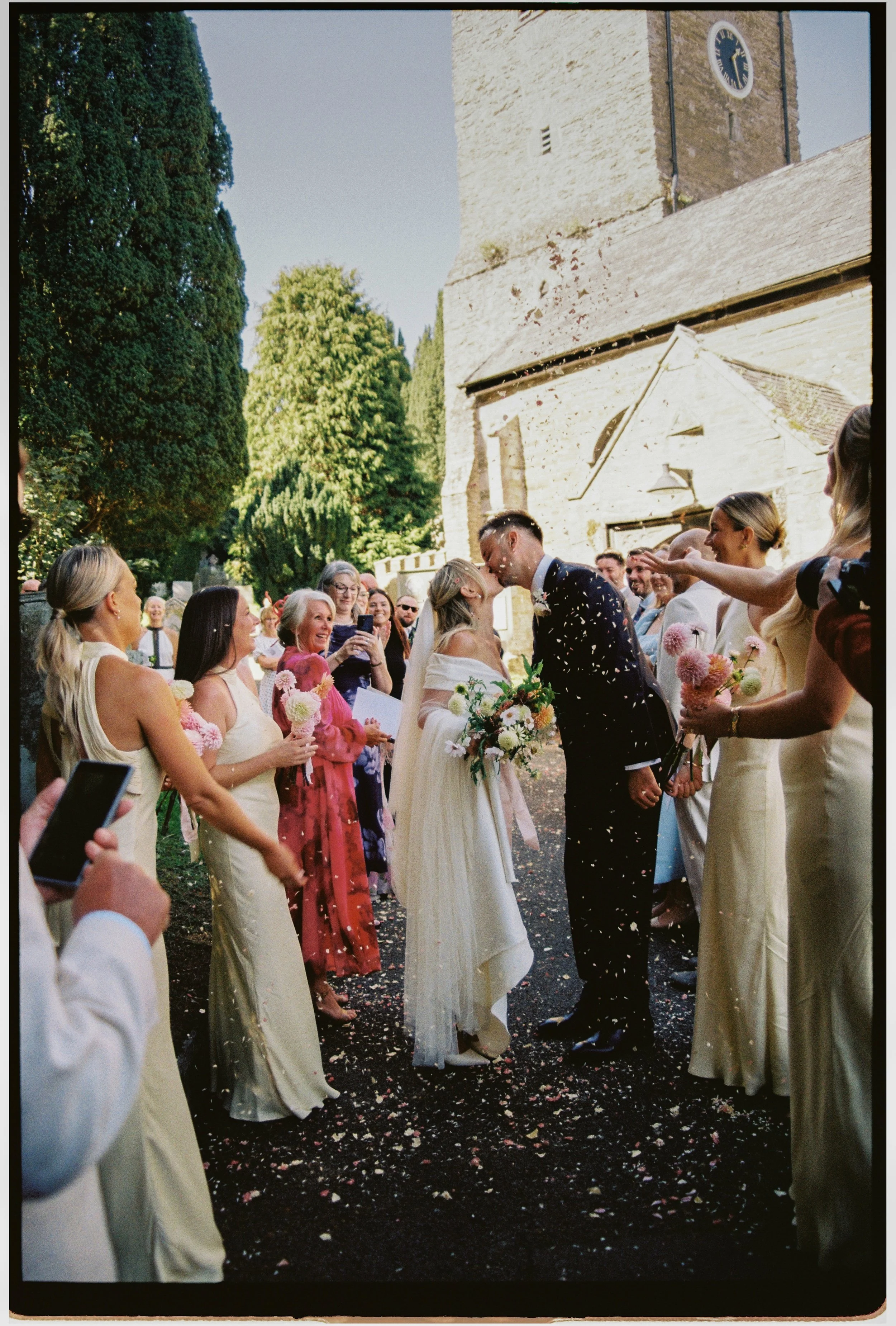 A bride and groom sharing a kiss during their outdoor wedding ceremony, surrounded by bridesmaids and guests, with a church building in the background and flower petals on the ground.
