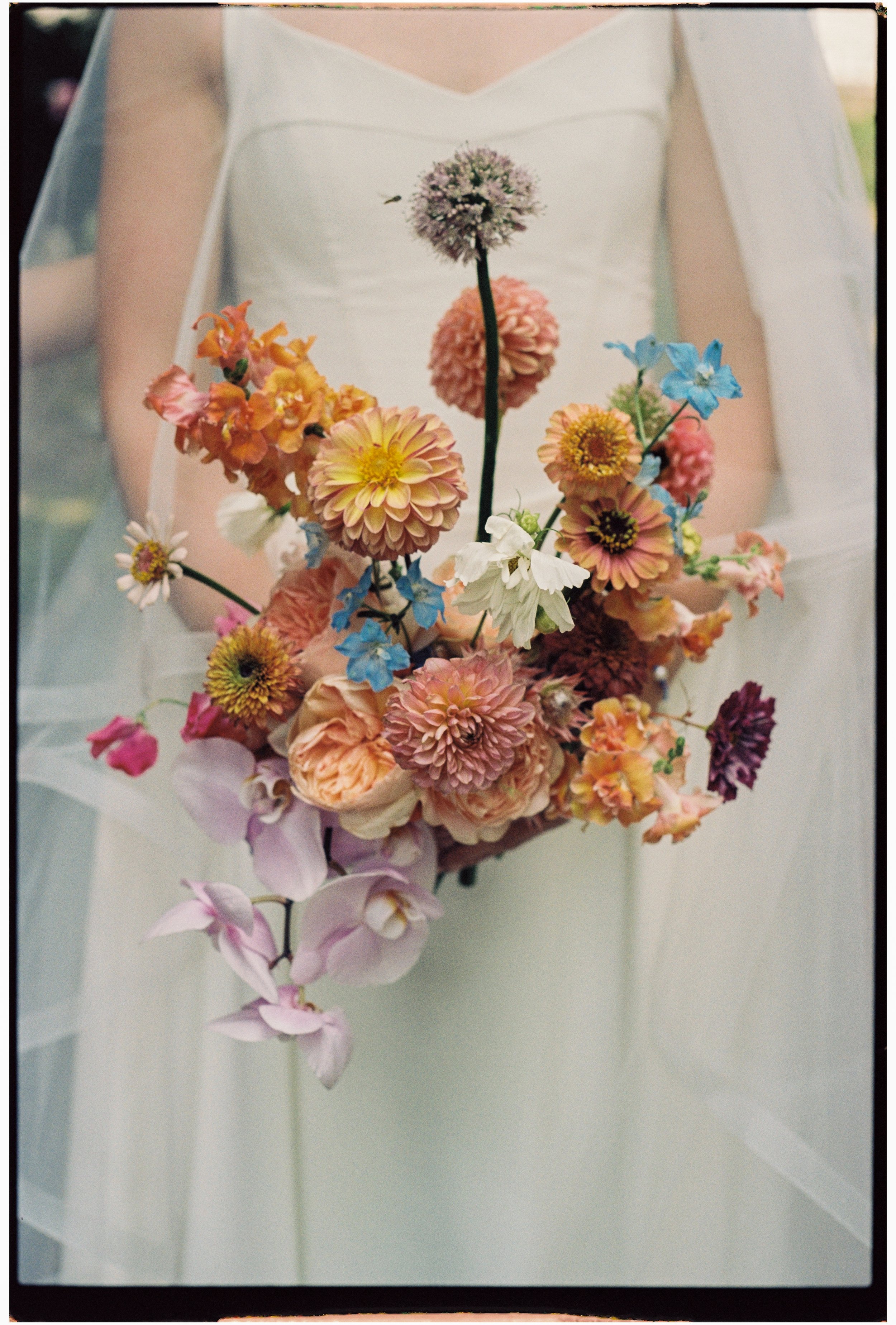 Close-up of a bride holding a colorful bouquet of various flowers, including pink, orange, white, and blue blooms, in a wedding dress with lace details on the sleeves.