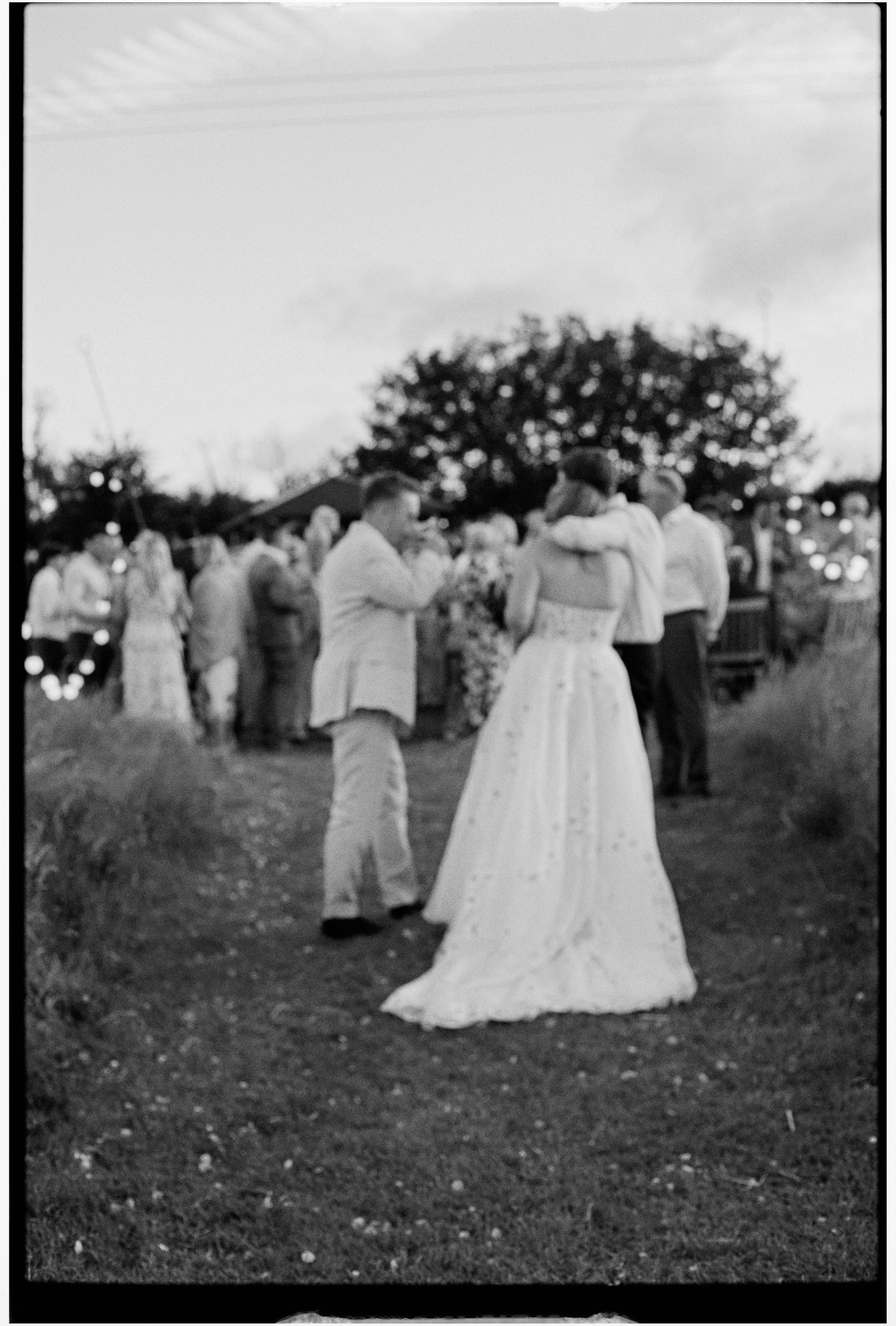 A black-and-white photo of a wedding ceremony outdoors at dusk, featuring a bride and groom holding hands, surrounded by guests with a large tree in the background.