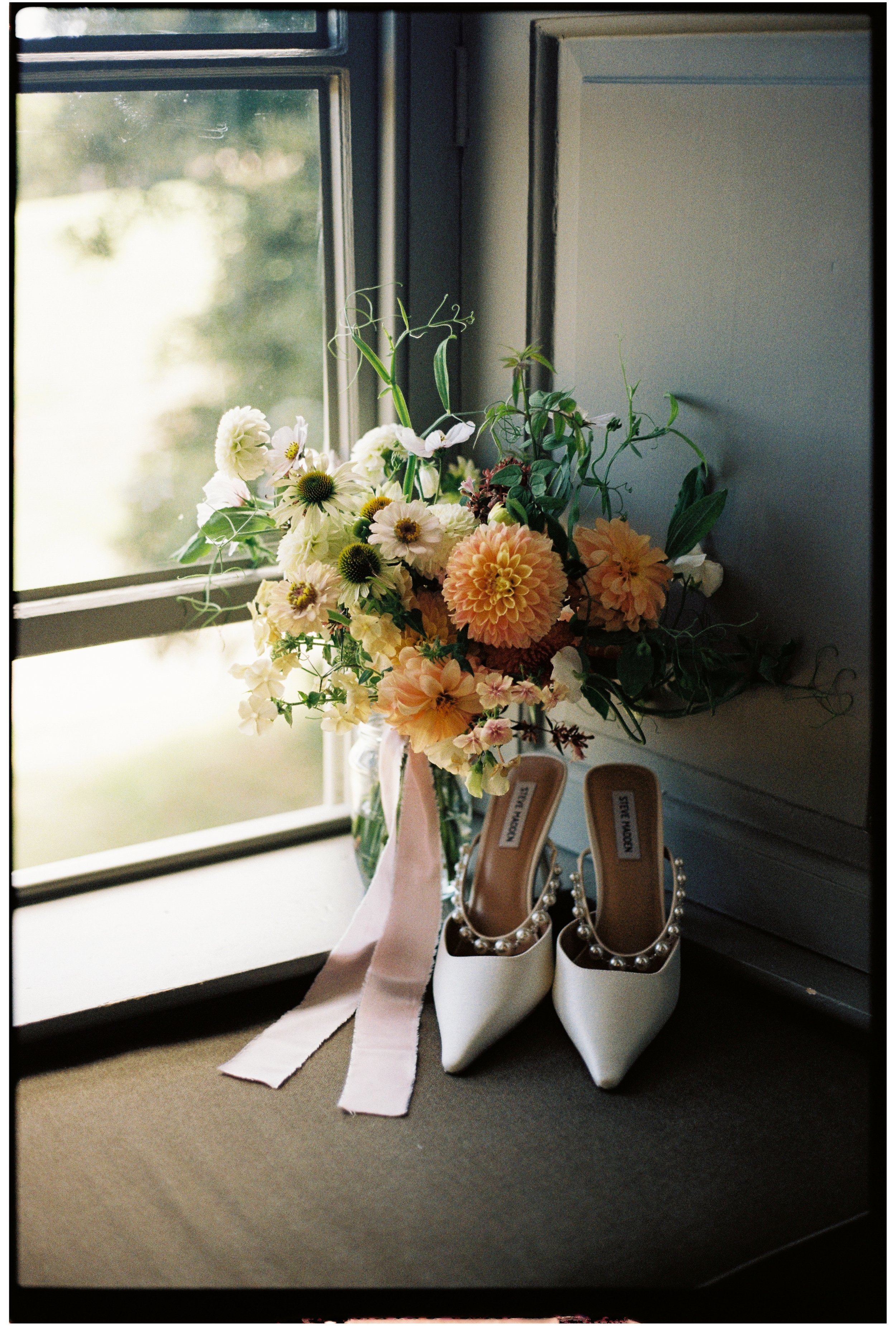 A floral bouquet with white, peach, and green flowers next to a pair of white high heel shoes adorned with pearls, all placed on a chest of drawers by a window at The Pig Hotel