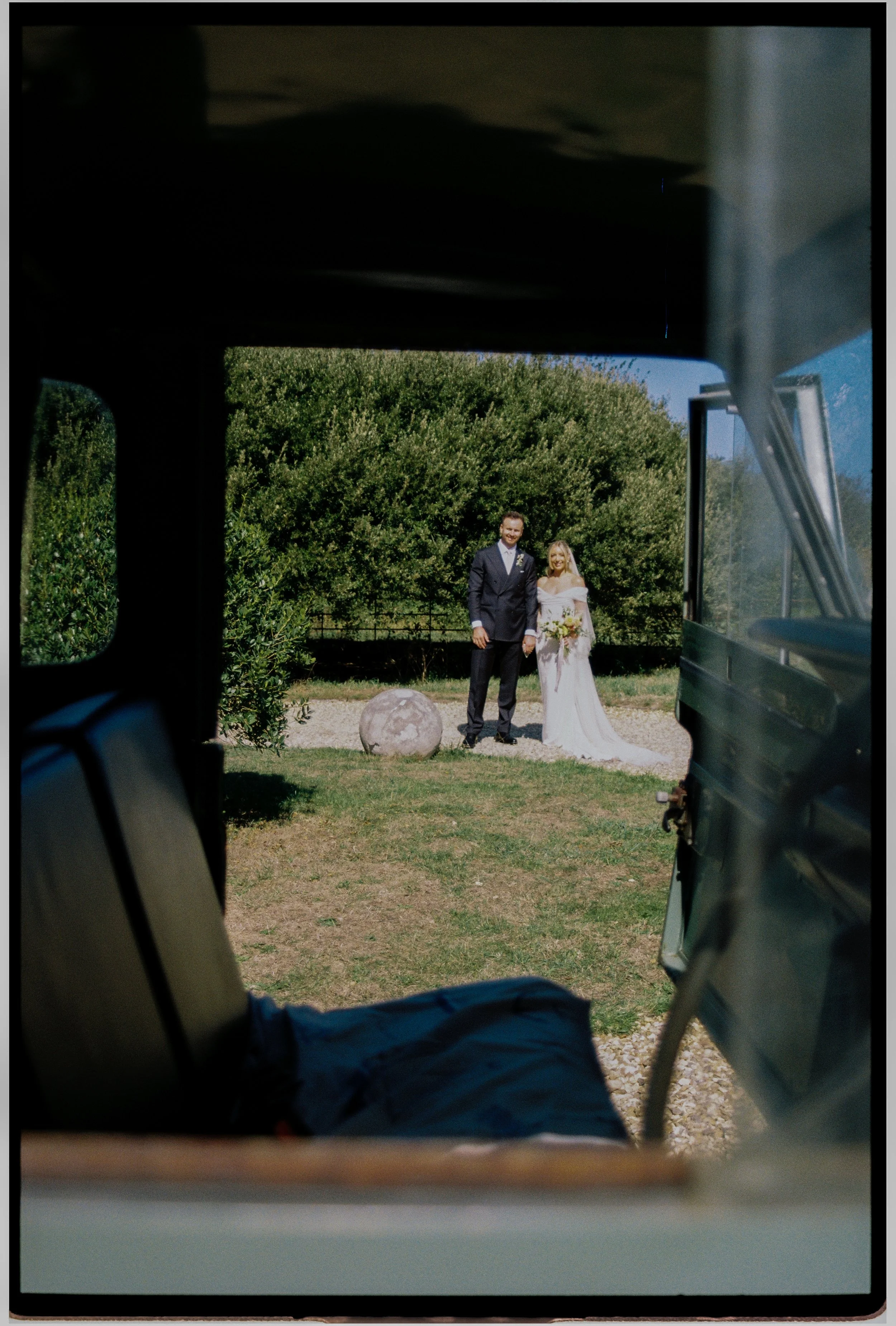 A bride and groom standing outdoors on a sunny day, framed by the opening of a vehicle, with greenery and trees in the background.