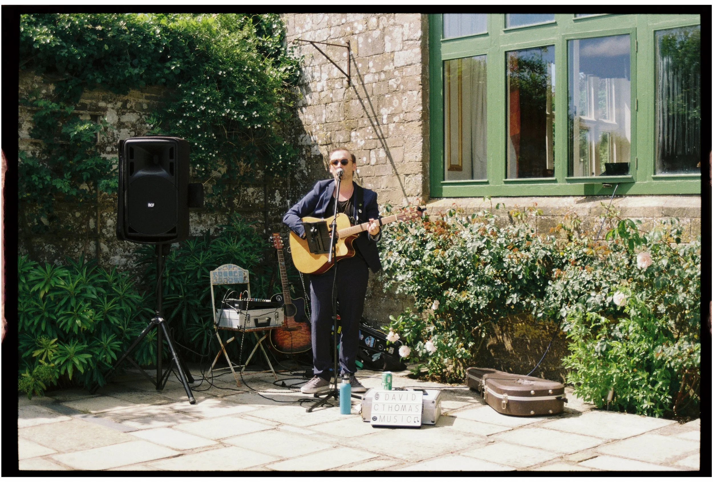 A musician is performing outdoors on a sidewalk, playing an acoustic guitar and singing into a microphone, surrounded by greenery and flowers, with a stone building and large window in the background.