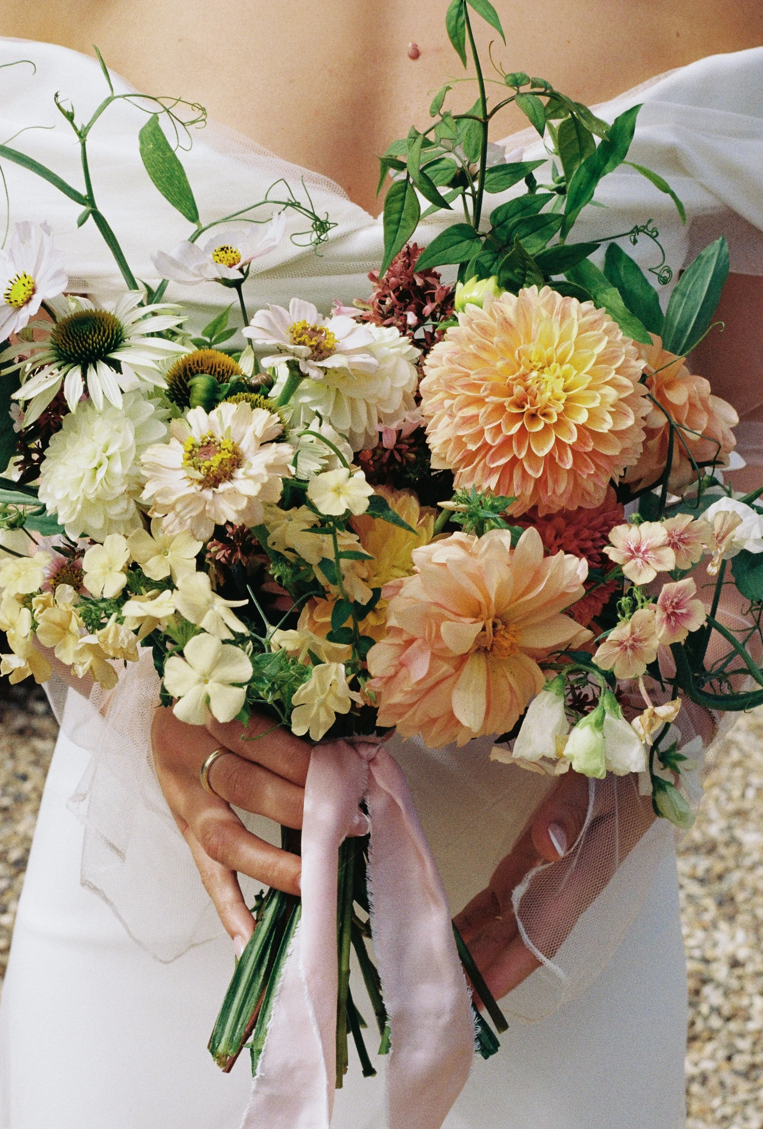 Person holding a colorful bouquet of flowers, including dahlias, daisies, and various greenery, wrapped with pink ribbon, in an outdoor setting.