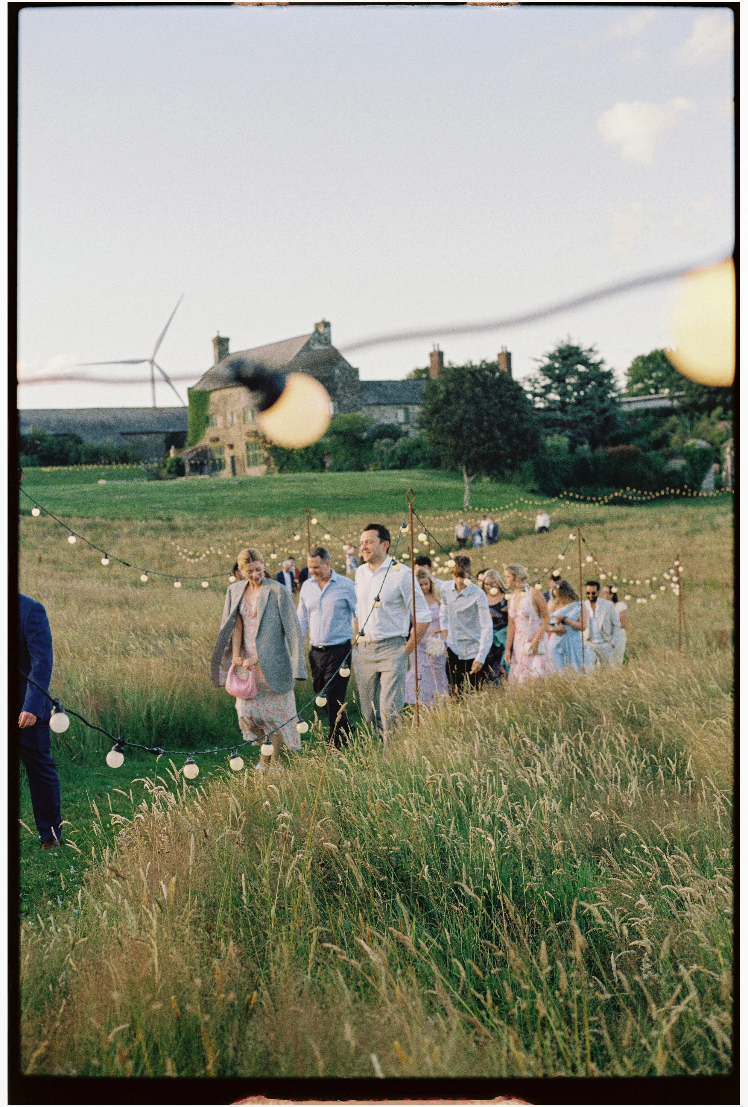 A group of people walking through a grassy field decorated with string lights, with a large house or castle and wind turbine in the background, during dusk.