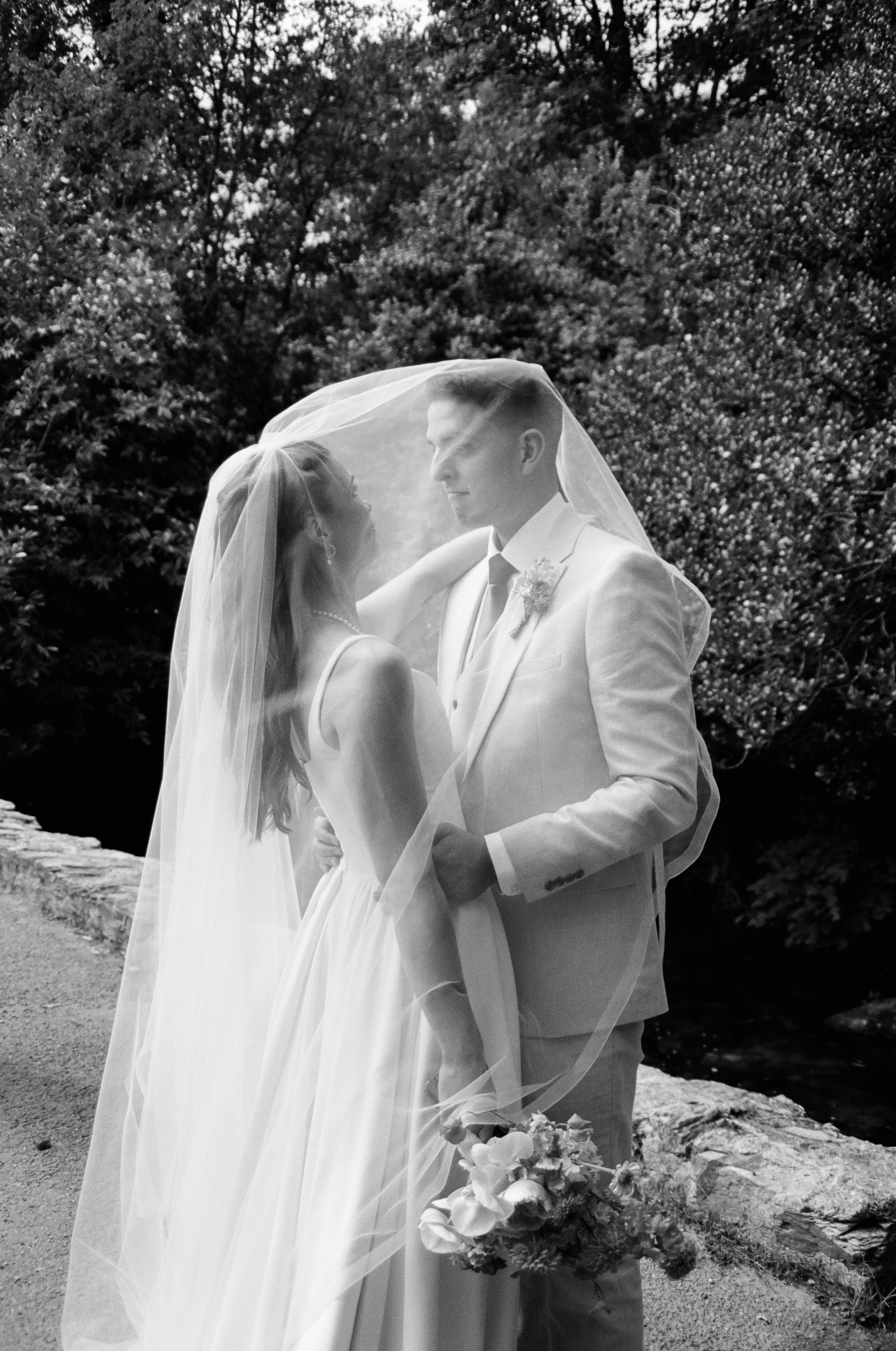A black and white photo of a bride and groom standing outdoors under a veil, with trees in the background. The bride is wearing a wedding dress and veil, and the groom is in a suit.