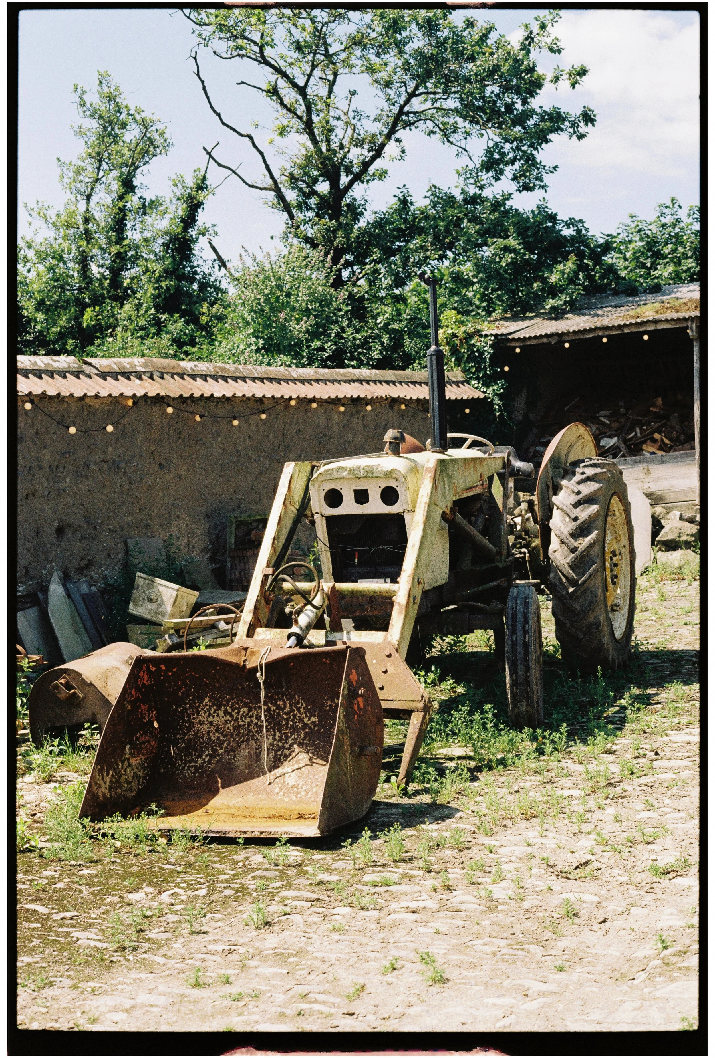 An old, rusty tractor with a front attachment is parked on a dirt surface with patches of grass. Behind it is a weathered building with a corrugated metal roof and a pile of wood. There are string lights hanging along the building. In the background,