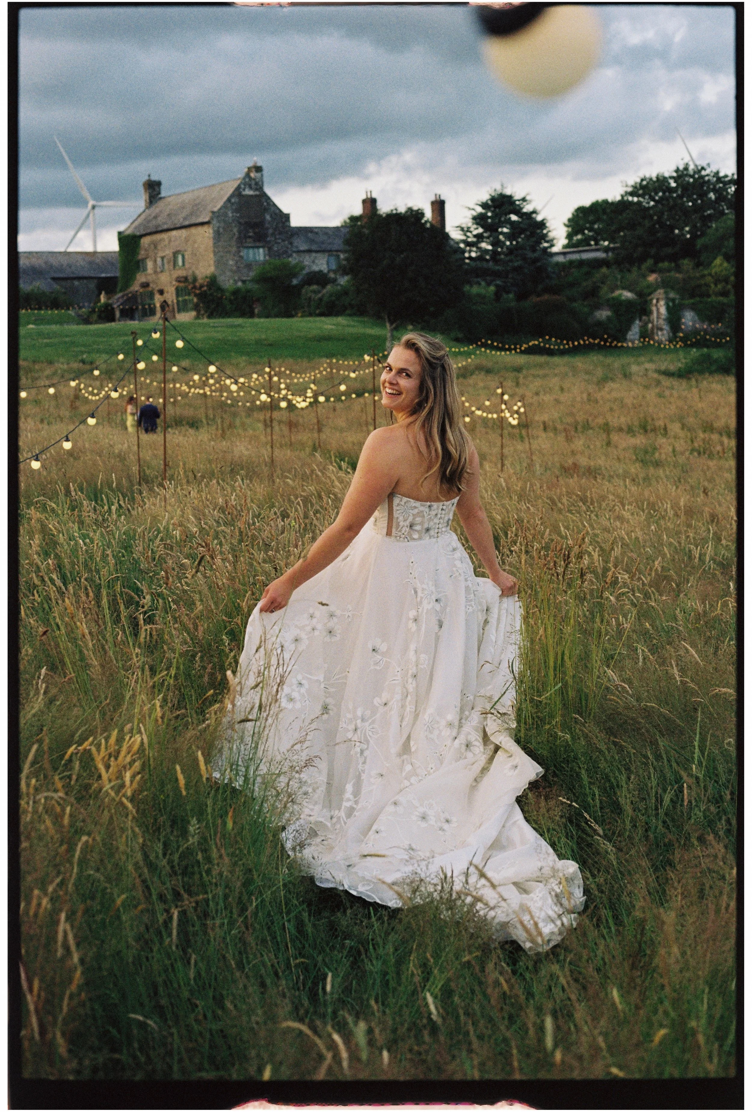 Bride in a white wedding dress standing in a grassy field with string lights in the background, under a cloudy sky.
