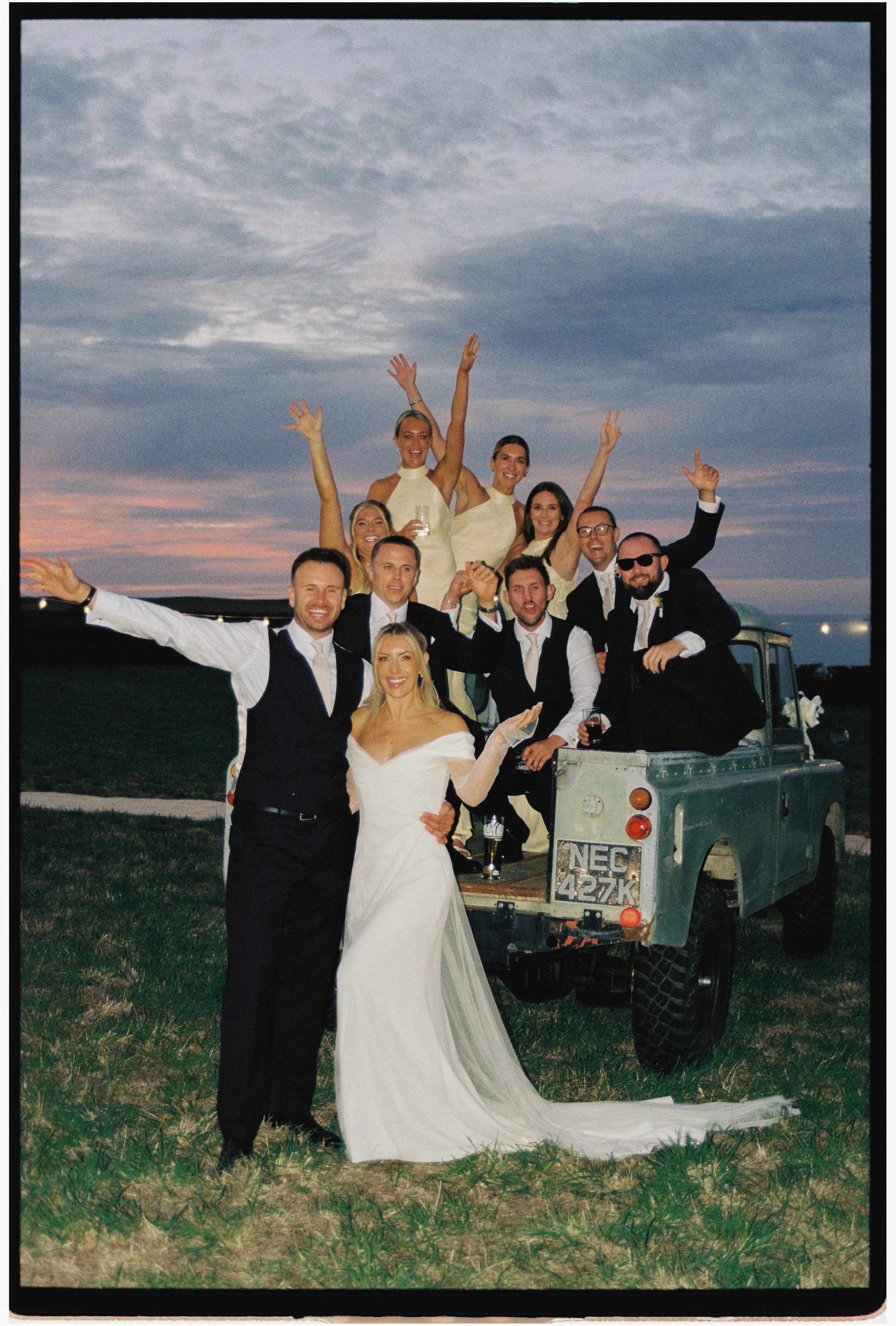 A newlywed couple poses in front of a vintage military vehicle with friends celebrating outdoors at sunset.