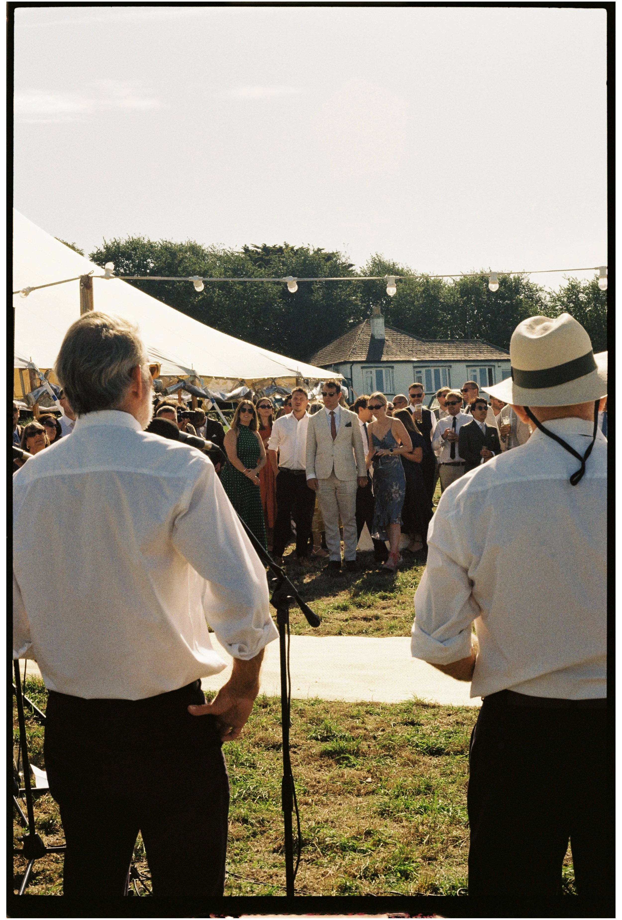 A group of people dressed in formal attire attending an outdoor wedding ceremony, with two men in white dress shirts and hats in the foreground, and the bride and groom with wedding guests in the background under a white canopy tent.