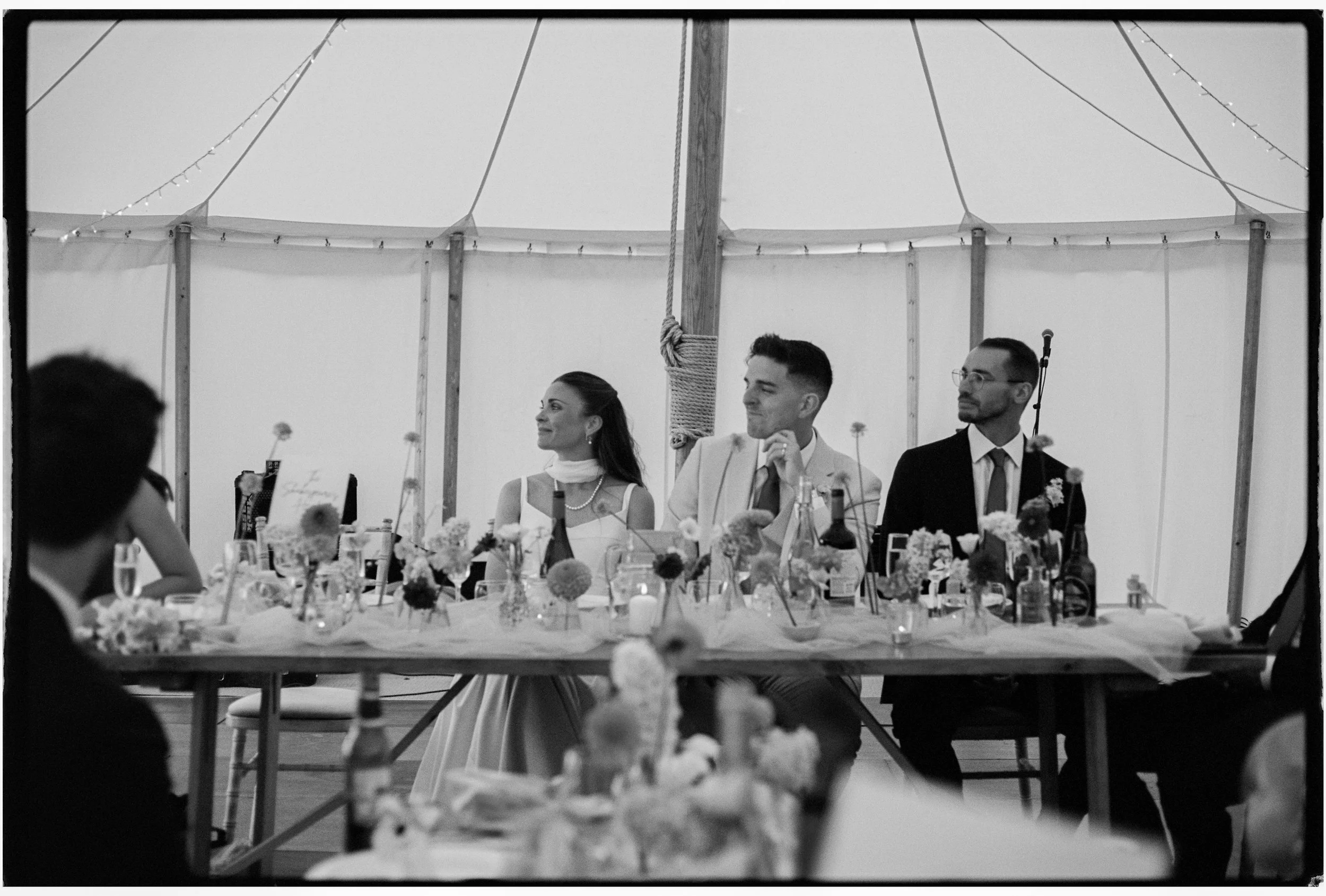 Group of people seated at a decorated dining table inside a large tent, with floral centerpieces and string lights overhead.