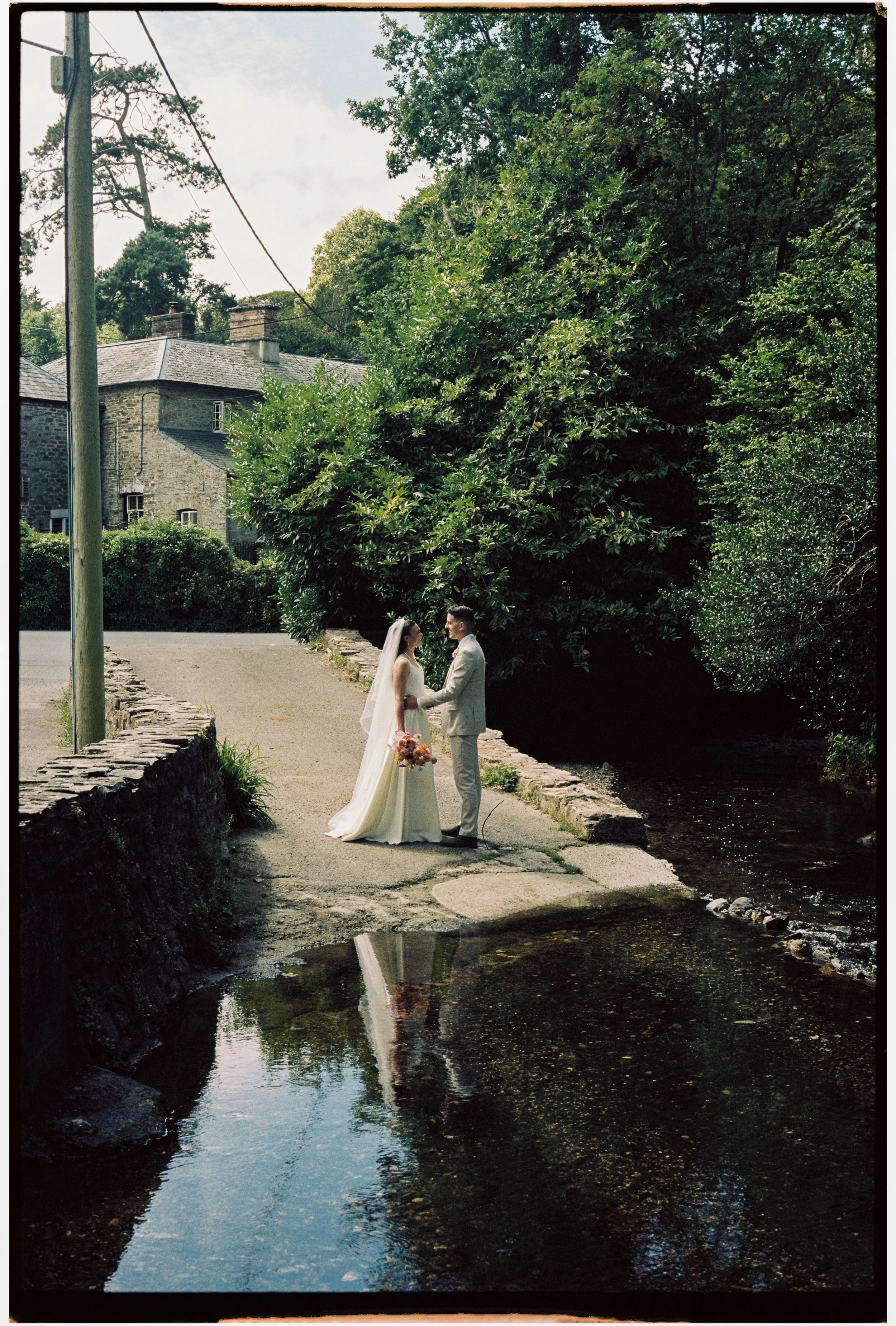 A bride and groom holding hands, standing on a small stone bridge over a creek in a lush green park with trees and a stone house in the background.