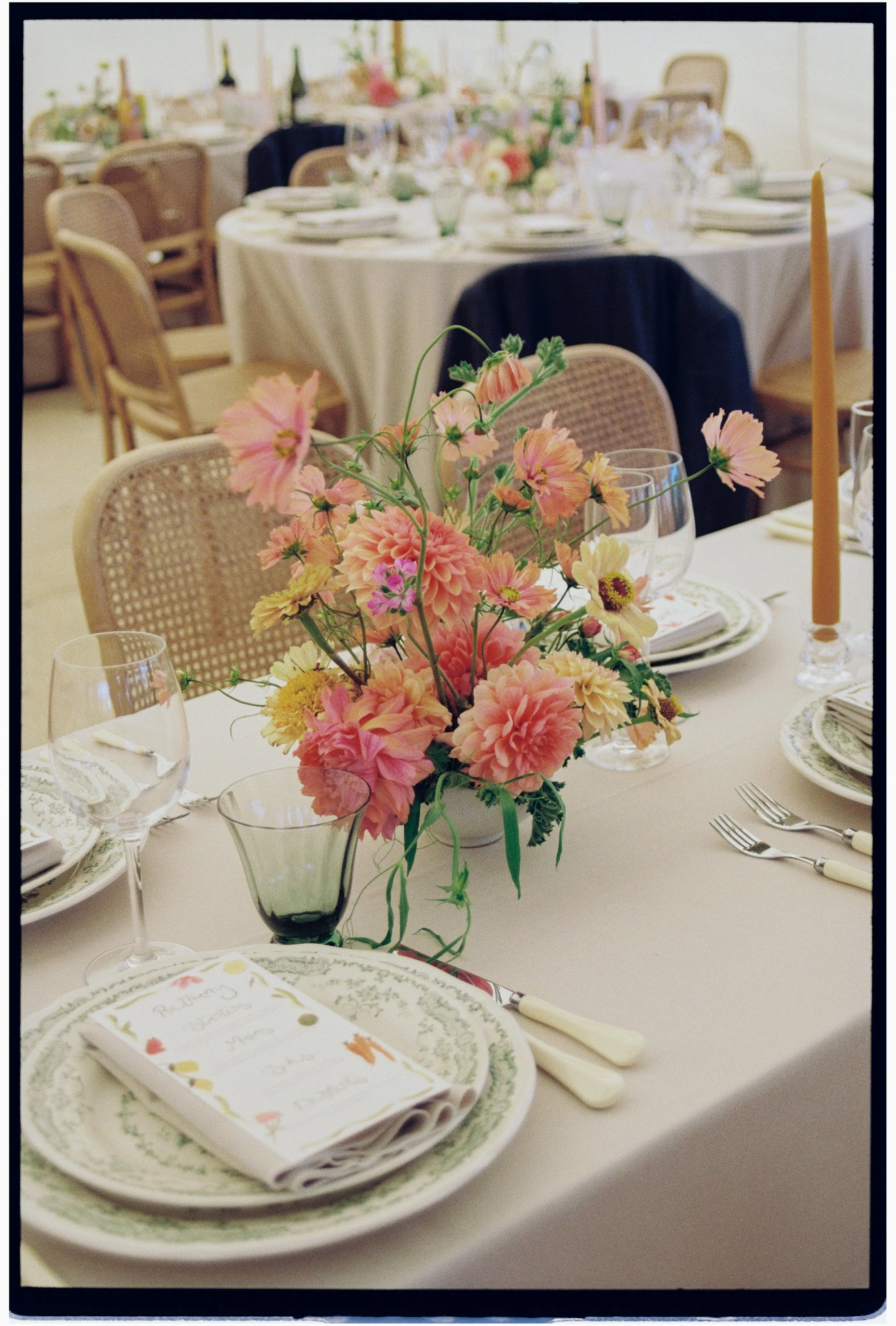 A close-up of a table setting for a formal event with a floral centerpiece, plates, napkins, silverware, wine glasses, and a tall pink candle on a white tablecloth.