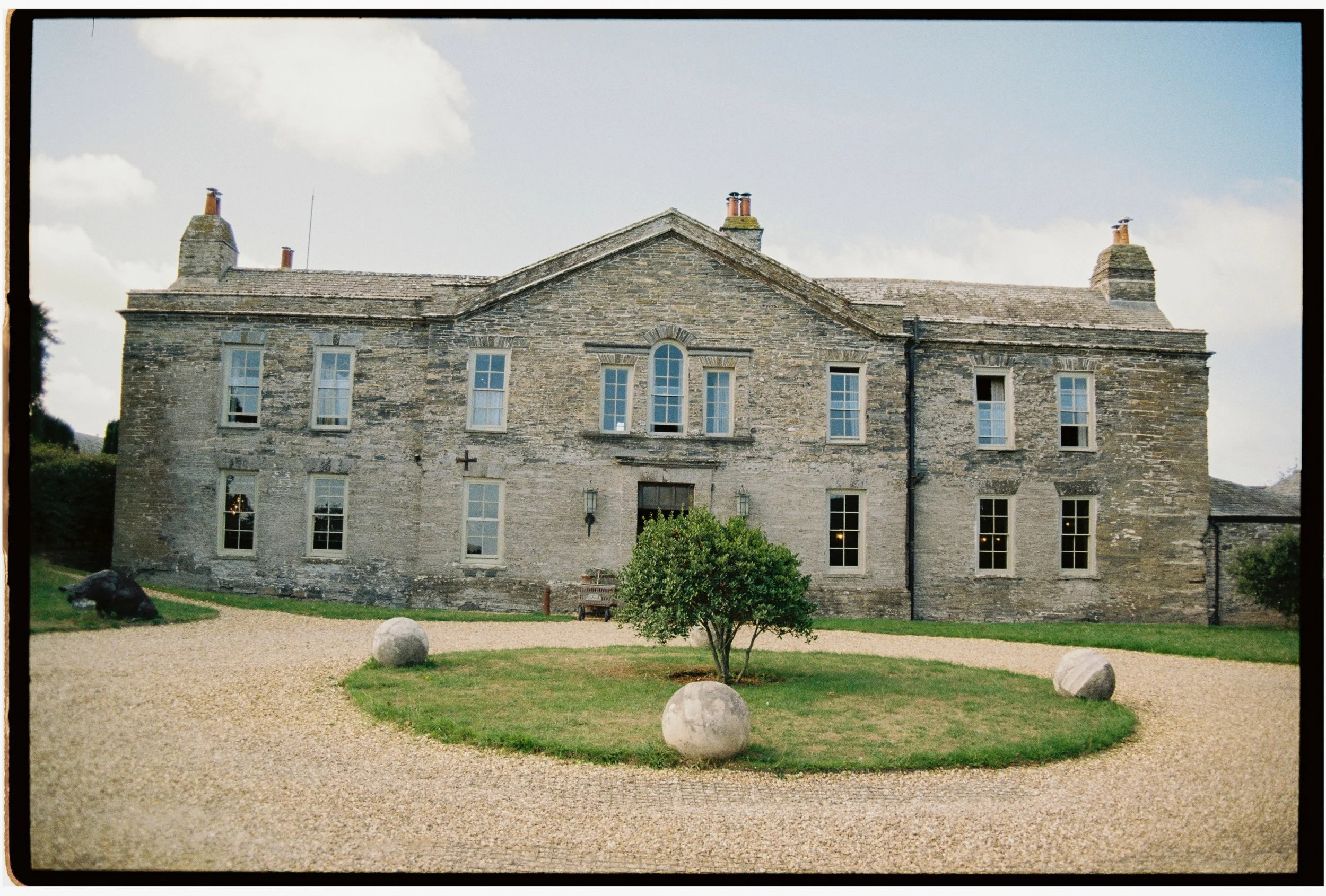 Historic stone building with multiple windows, chimneys, and a gravel courtyard with spherical stone decorations and a small tree.