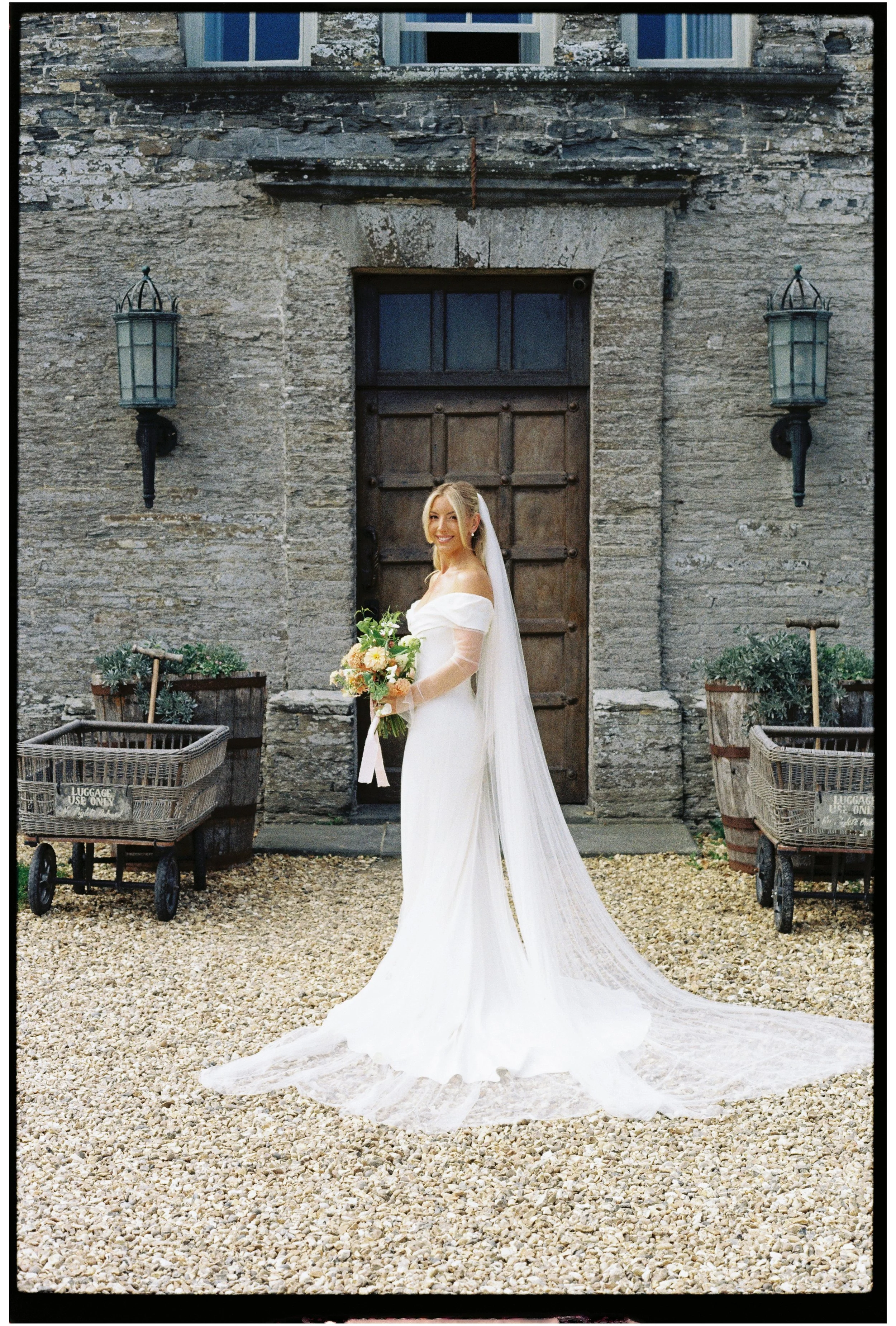 A bride in a white wedding dress holding a bouquet of flowers standing outside in front of the Pig Hotel Harlyn with wooden door and two lanterns, on a gravel ground for her wedding in Cornwall
