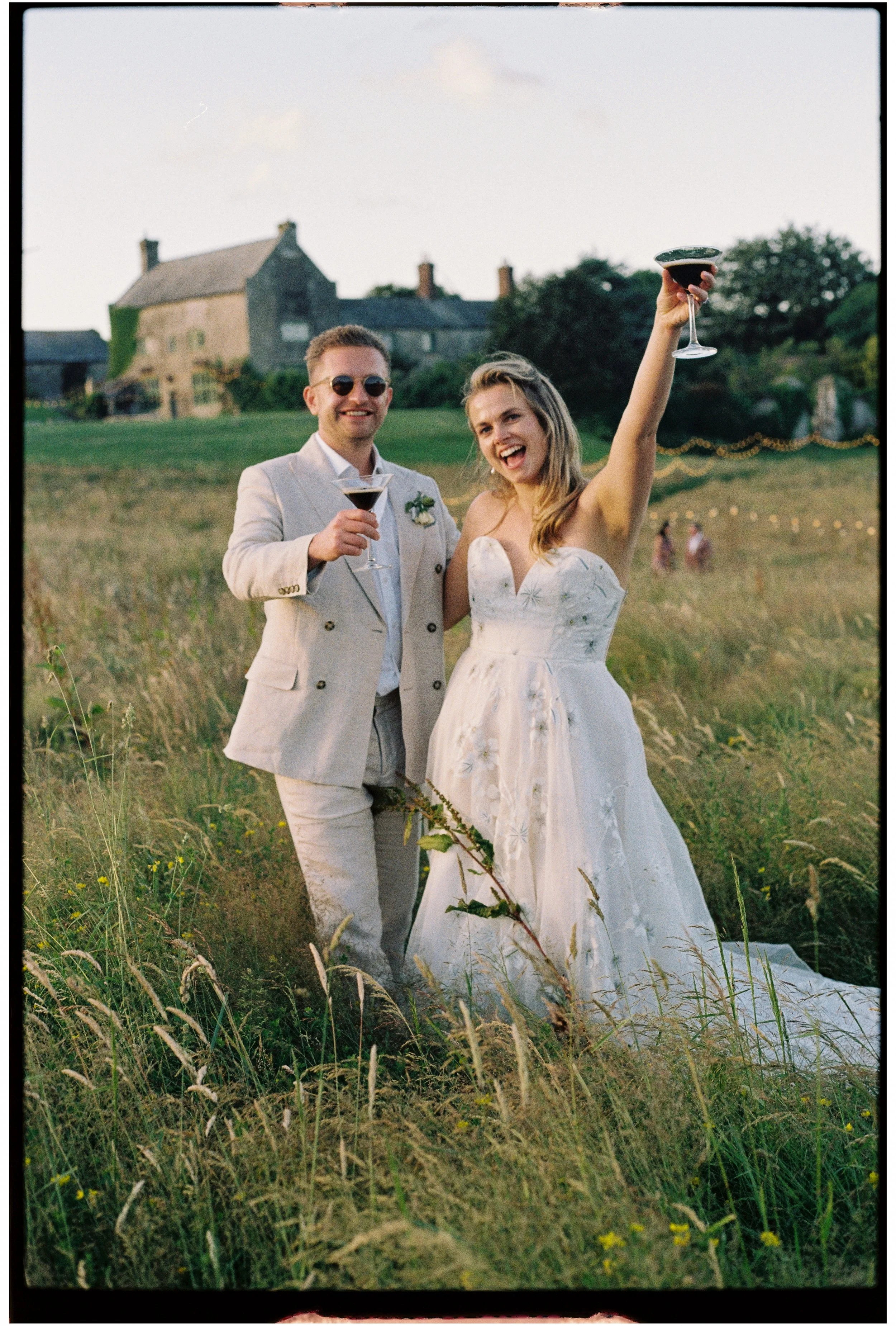 A couple dressed in wedding attire standing in a field of tall grass, holding glasses of wine and smiling, with a large house or barn in the background during daytime.