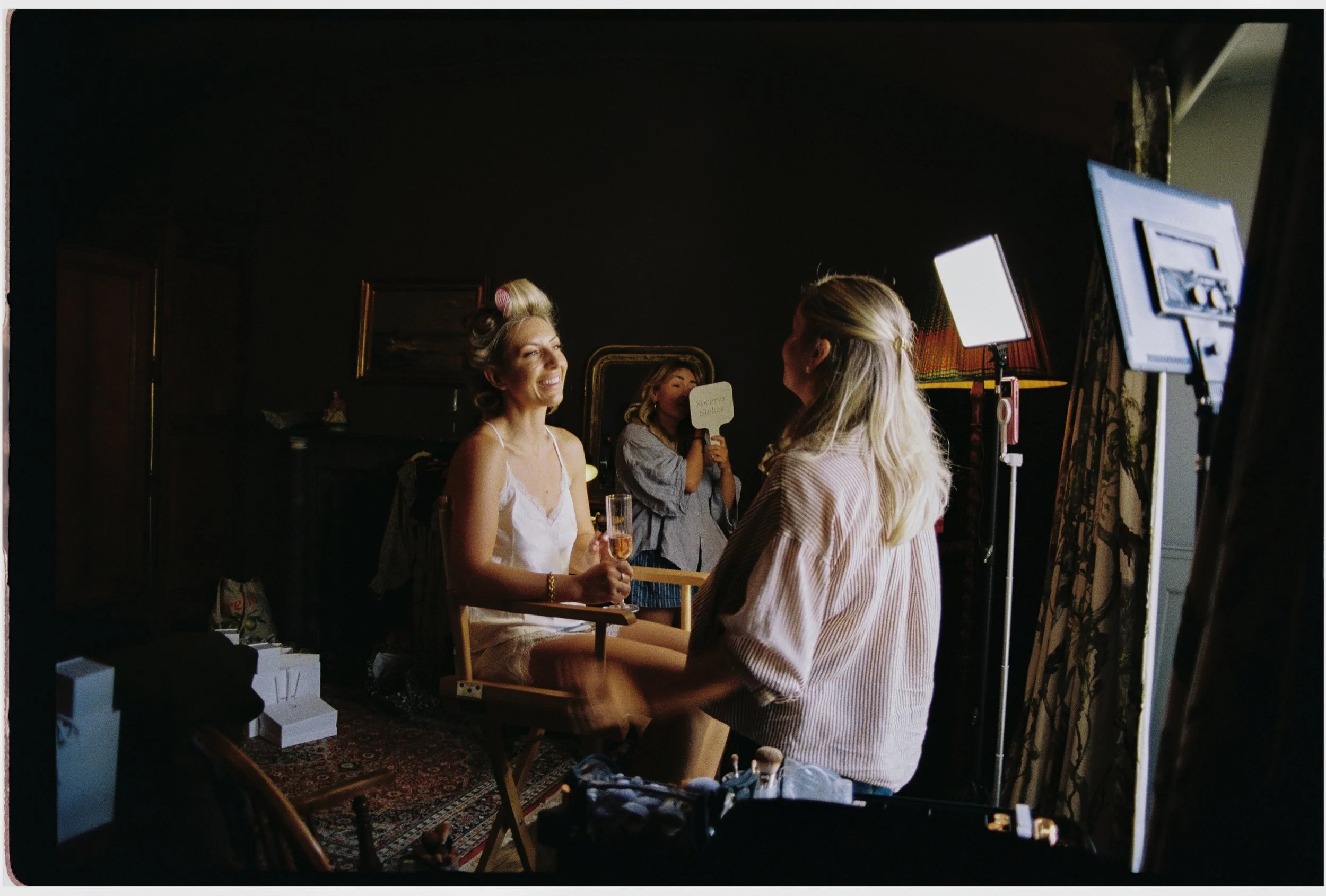 A woman with vintage hair rollers holding a glass of champagne is having her makeup done by Charlotte Albert at The Pig Hotel Harlyn while Soccora Hairstylist does her make up for the wedding in Cornwall